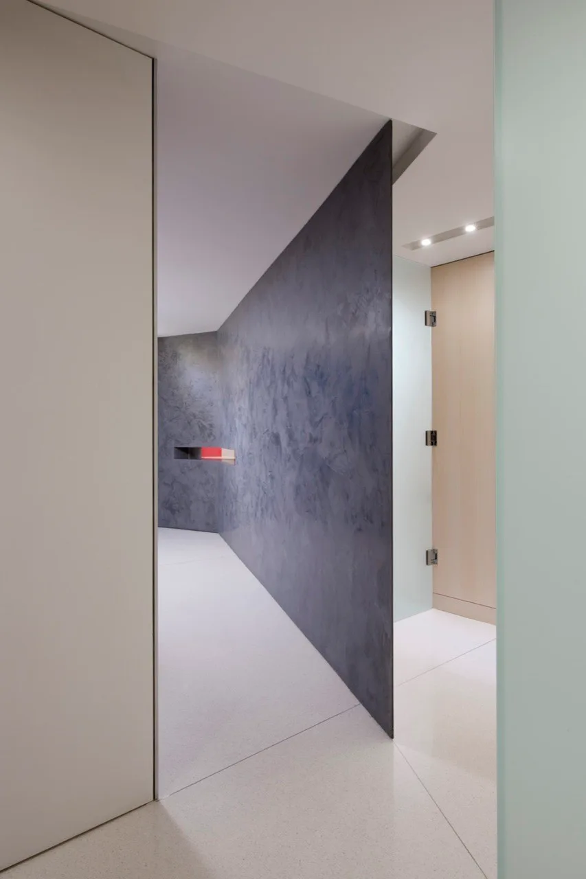 A minimalist hallway features an angled textured dark gray plaster wall, a light wood door, white floors, and recessed ceiling lighting.