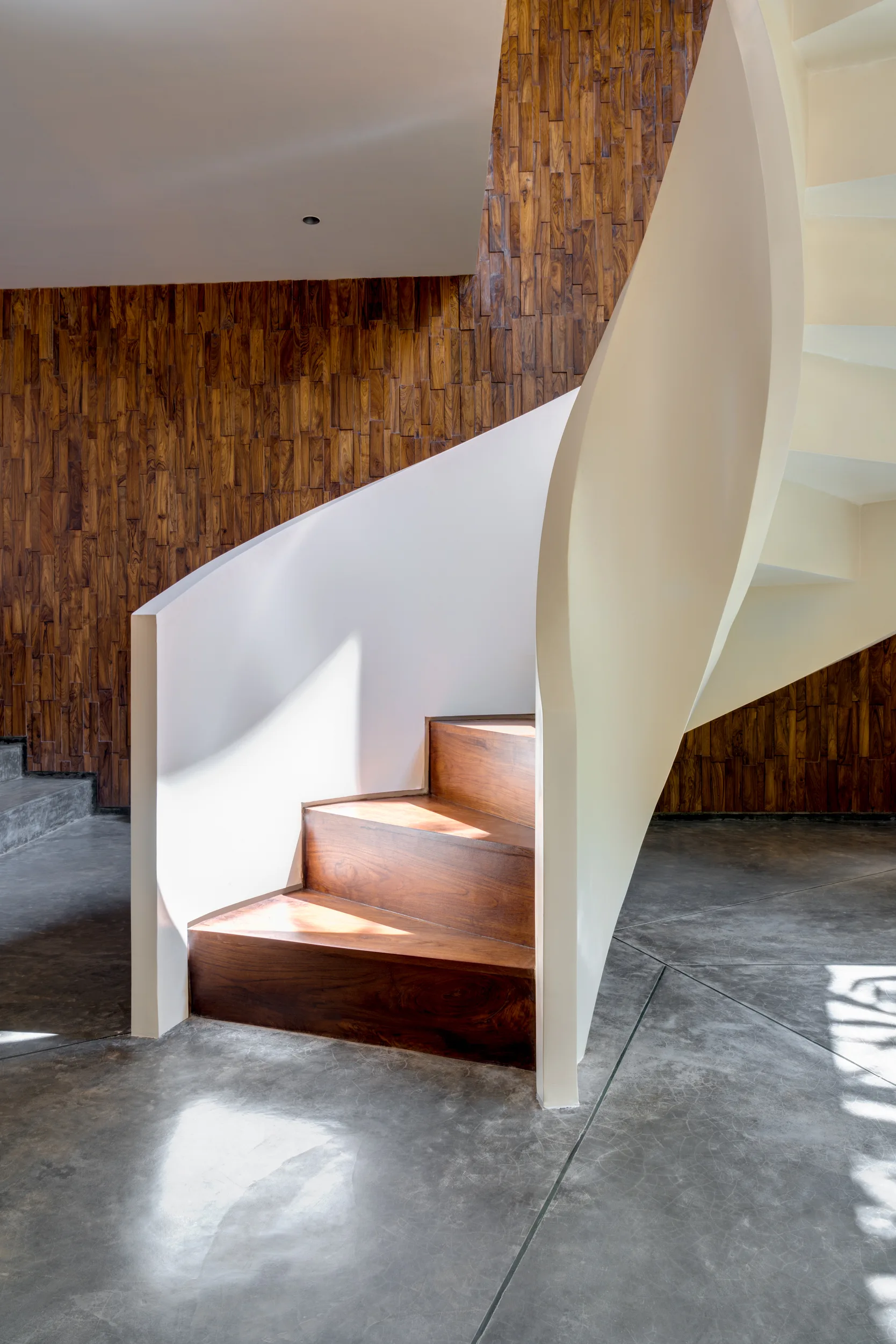 A modern curved staircase with dark wood steps and a solid white railing stands on concrete flooring in front of a textured wood-block accent wall.