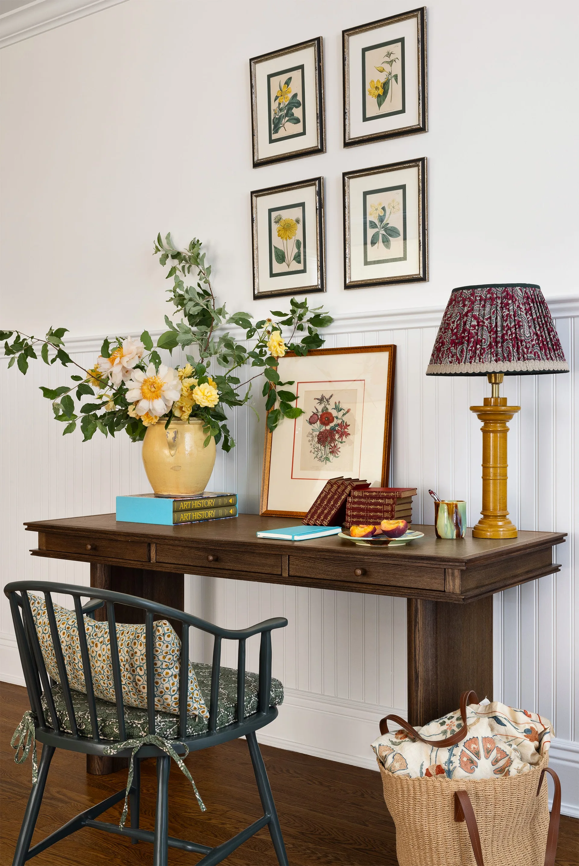 A dark wood writing desk set against white beadboard wainscoting features a patterned table lamp, botanical art prints, and a spindle-back chair.