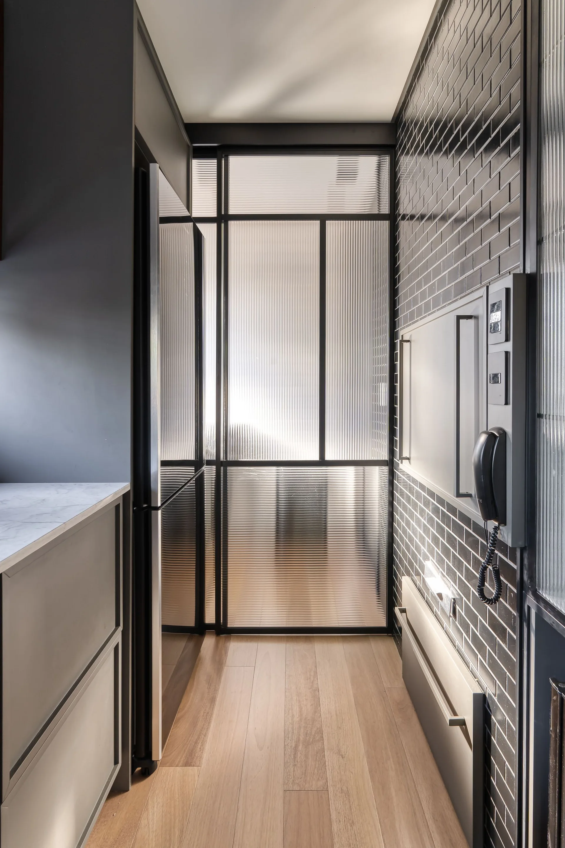 A narrow hallway features a ribbed glass partition with a black frame, light wood flooring, and a wall clad in glossy black subway tiles.