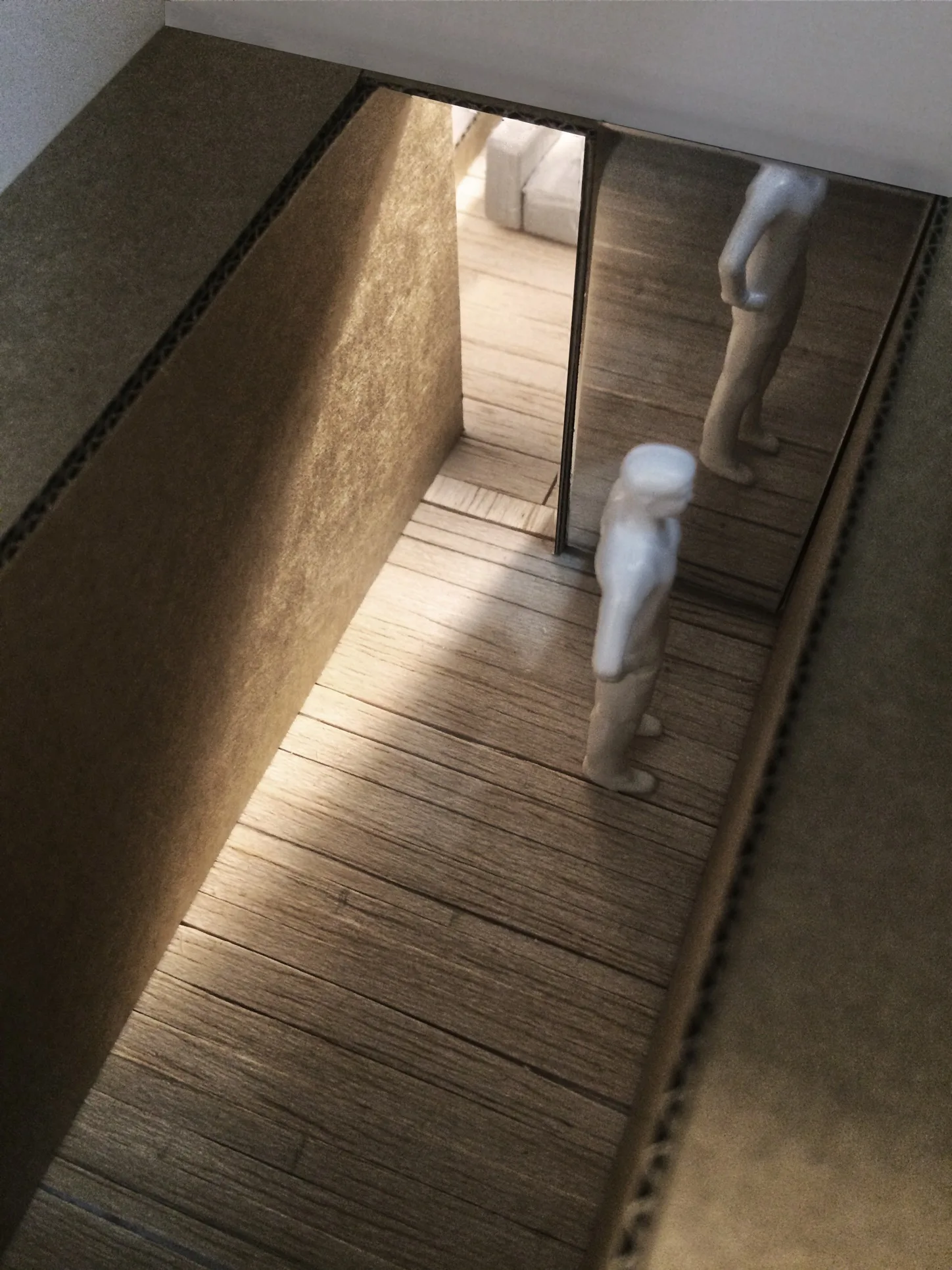 An architectural scale model features cardboard walls, miniature white figurines, wood-look plank flooring, and a mirrored panel at the end of a narrow hallway.