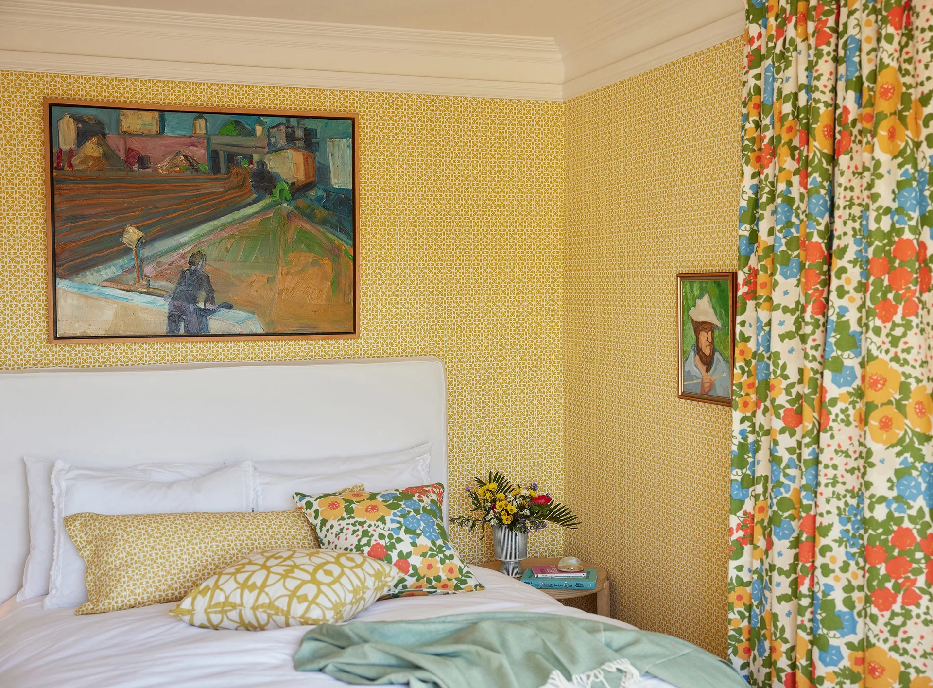 A bedroom features yellow geometric wallpaper, a white headboard, mixed pattern pillows, and colorful floral curtains.