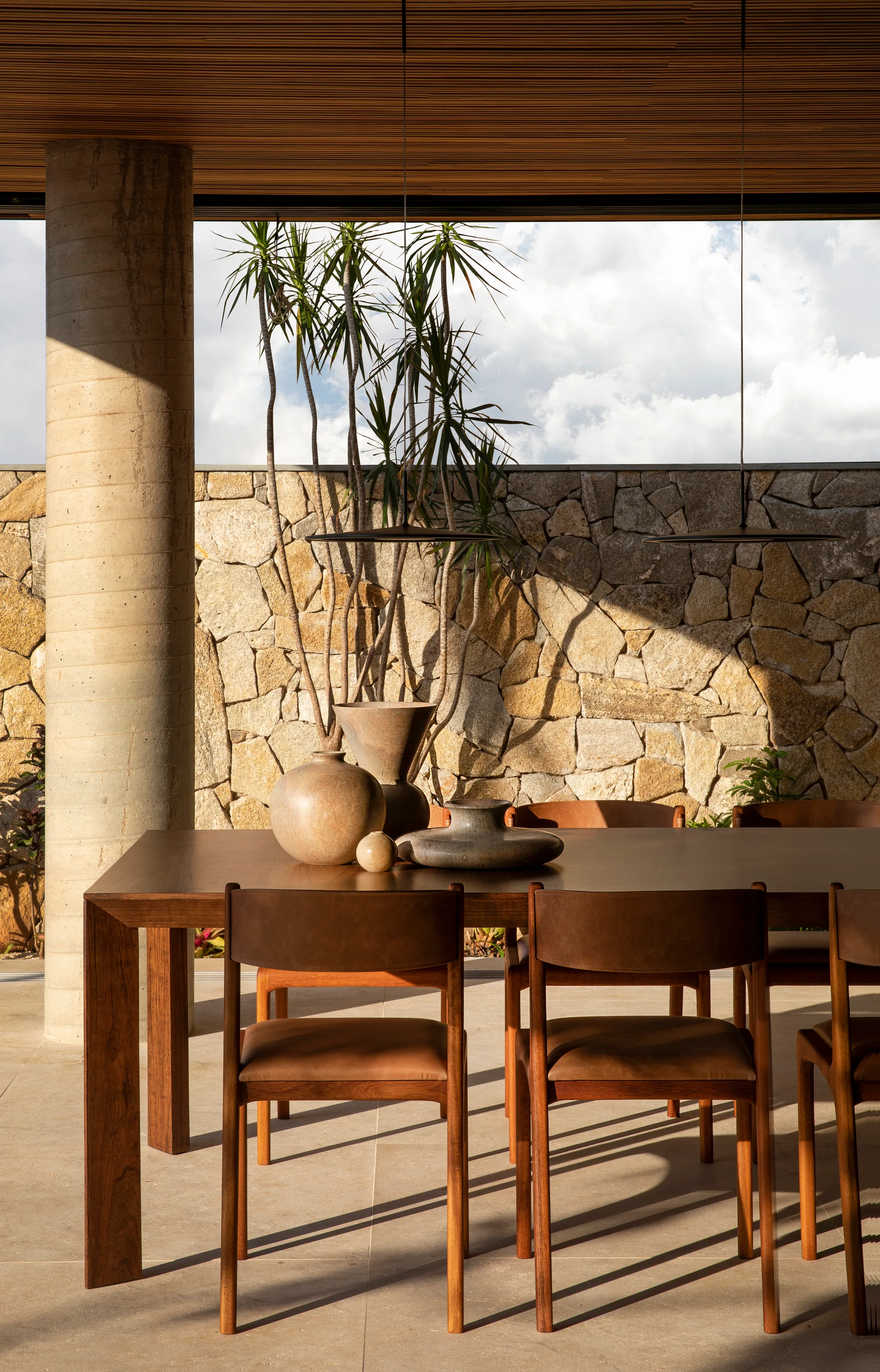 A wooden dining set with leather chairs sits on a stone patio beneath a slatted ceiling, backed by a textured stone wall.