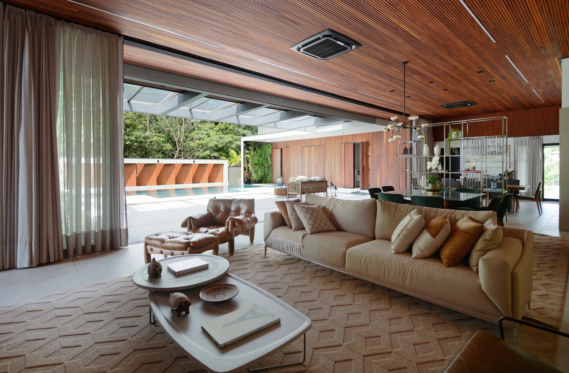 A modern open-plan living room featuring a tan sofa, nested coffee tables, a wood slatted ceiling, and large glass doors opening to a pool.
