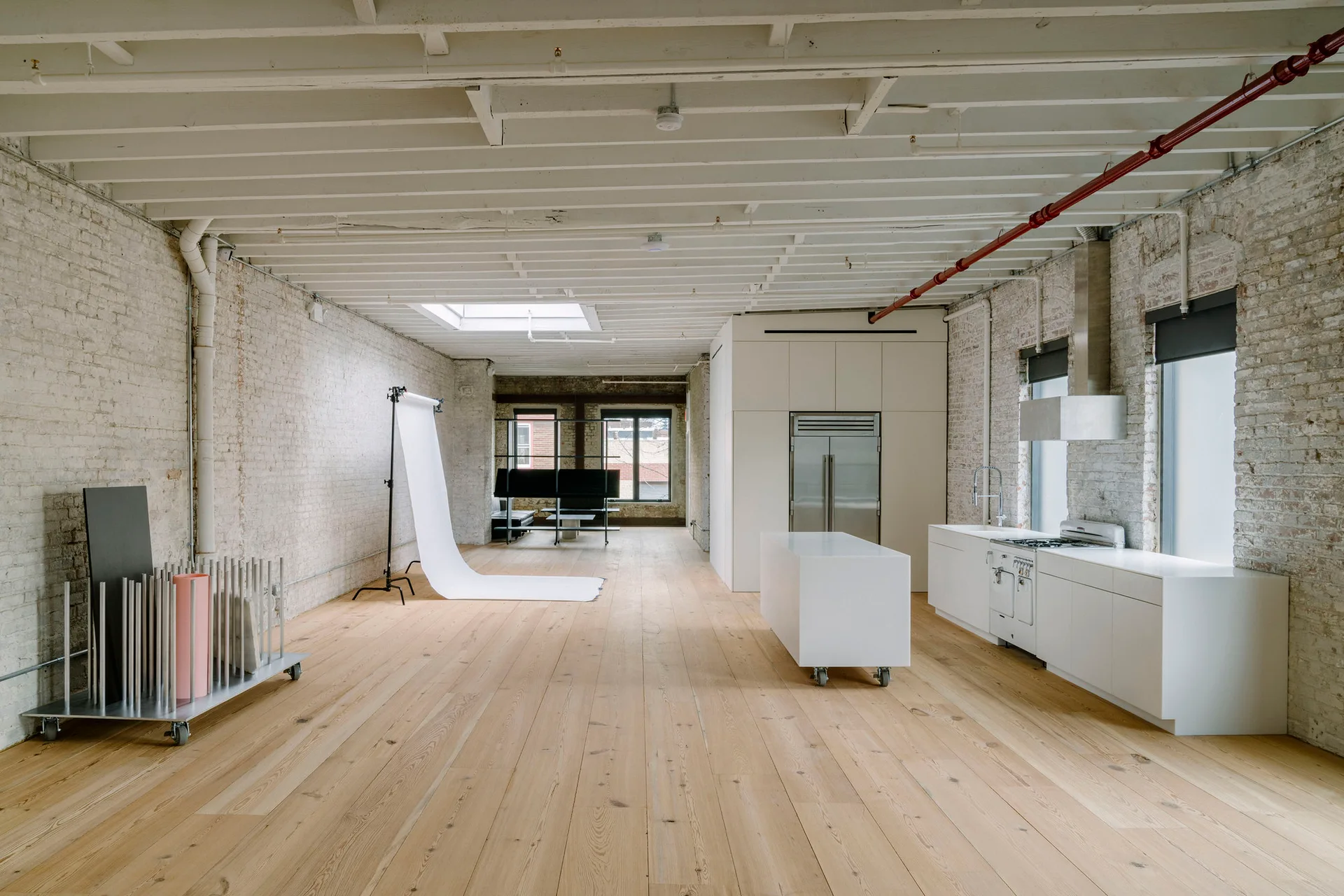 A spacious open industrial loft features a minimalist white kitchen with a mobile island on wheels, whitewashed brick, and light wood flooring.