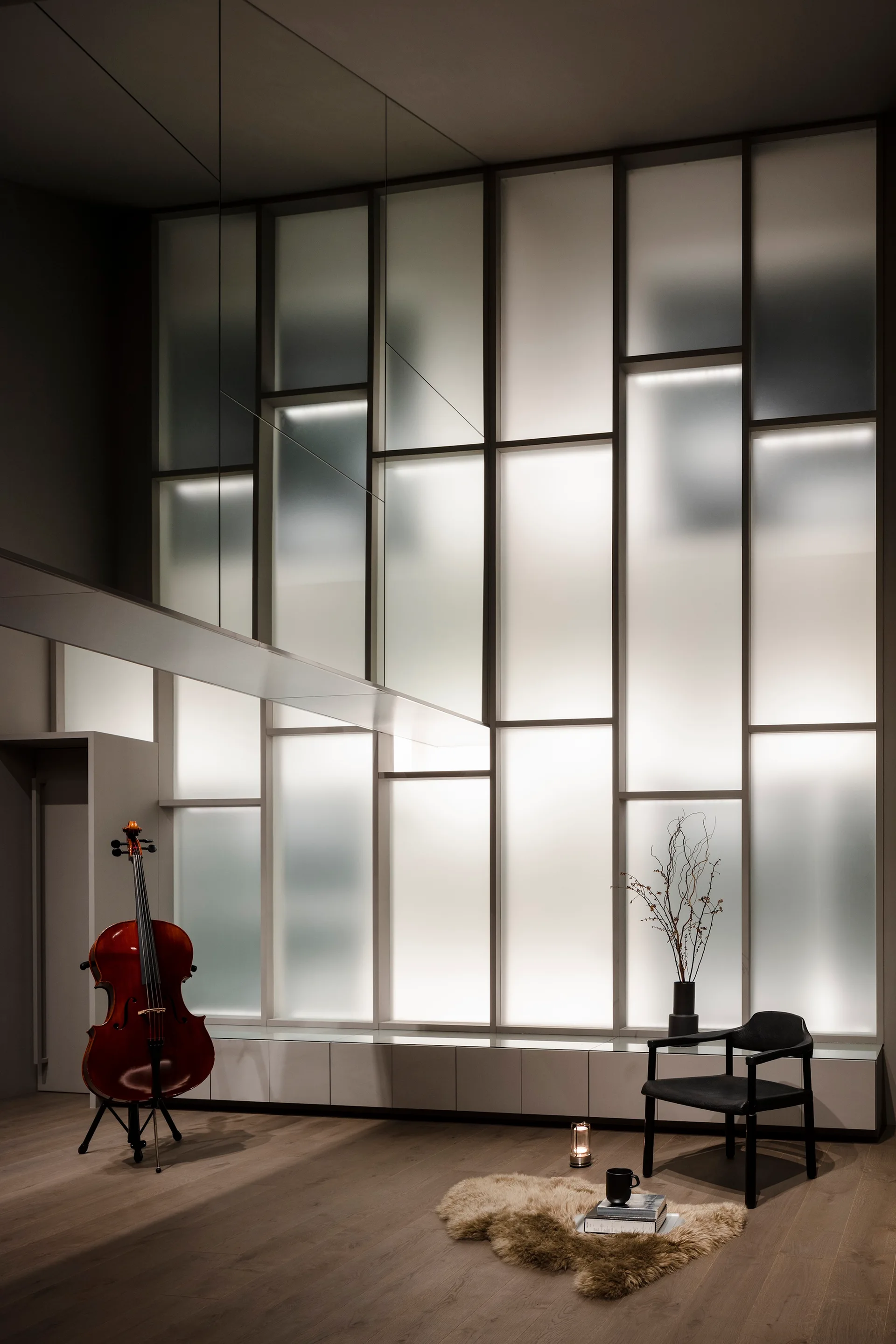 A minimalist room features a large backlit frosted glass grid wall, a cello on a stand, a black armchair, and a small fur rug on a wooden floor.
