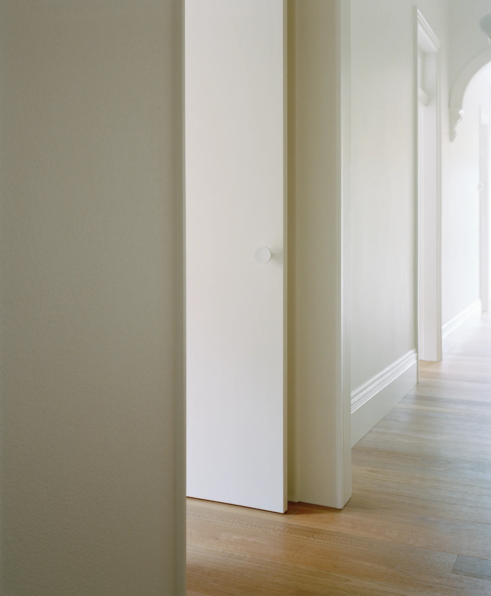 A white minimalist door with a round knob stands slightly open in a bright hallway featuring light wood flooring and traditional trim.