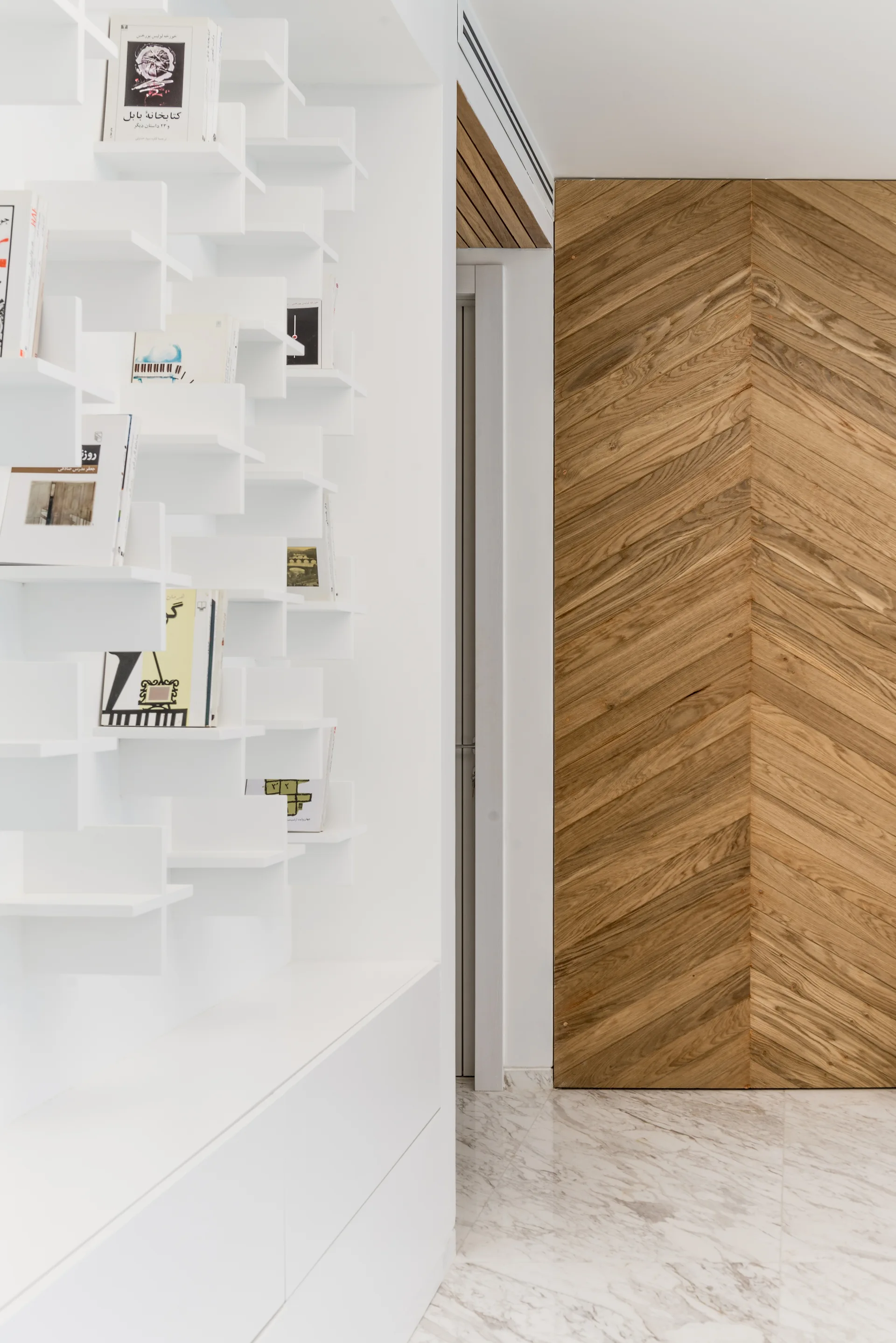 A minimalist hallway features a white geometric wall bookshelf next to a full-height wood door with a herringbone pattern on marble flooring.