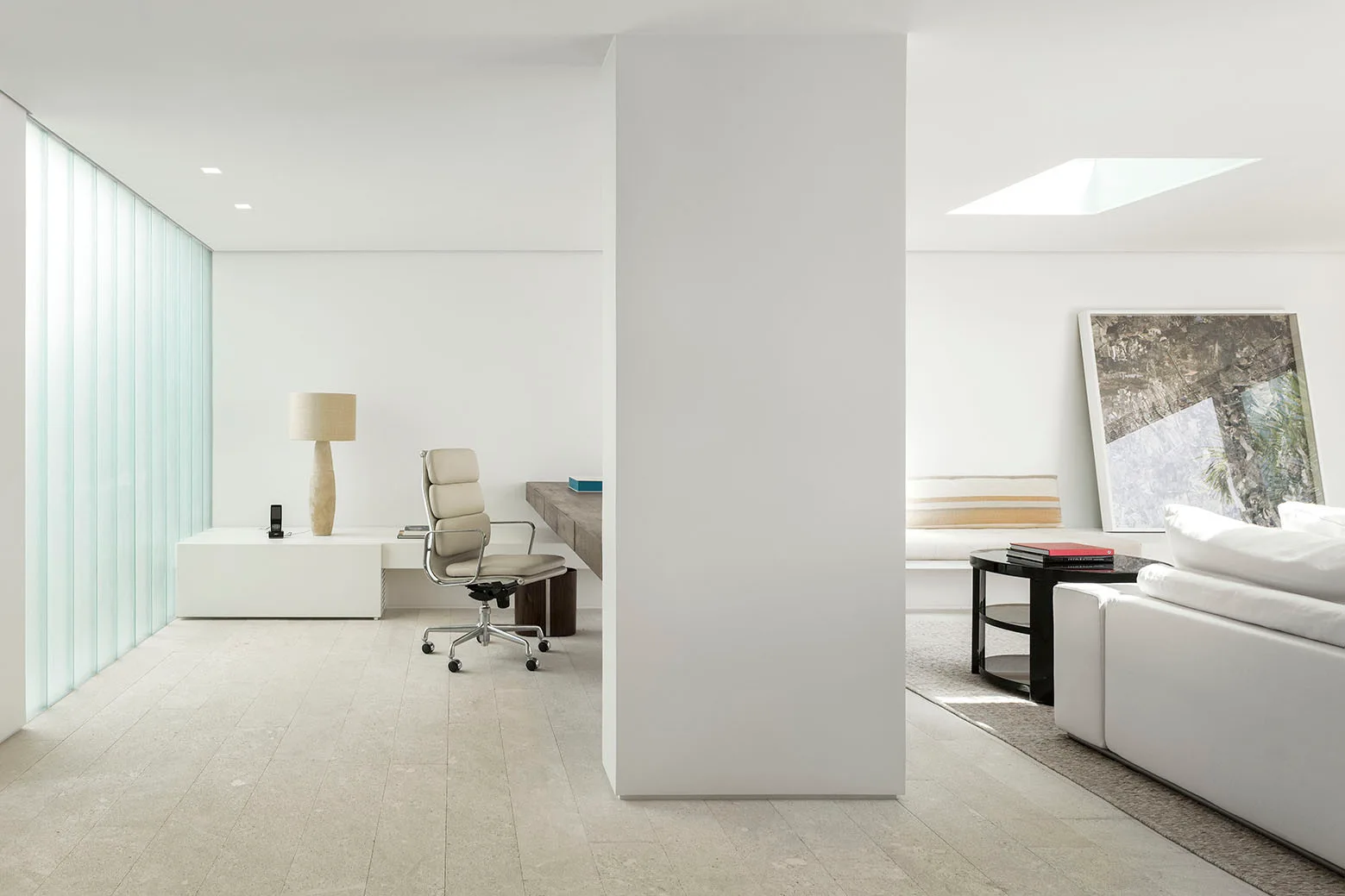 A minimalist living space featuring a central white column separating a home office with a floating wooden desk from a living room with a white sofa.