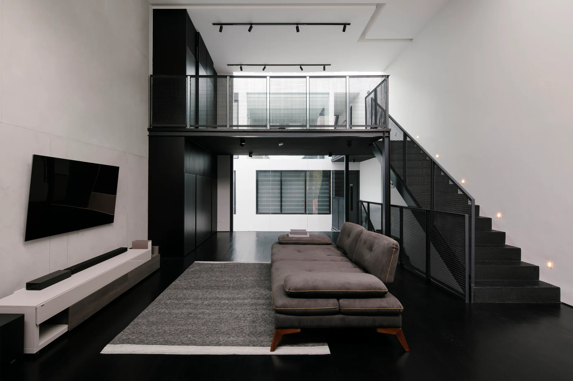 A minimalist living room features a dark grey sofa, black floors, and a black metal grid staircase leading to an upper mezzanine level.
