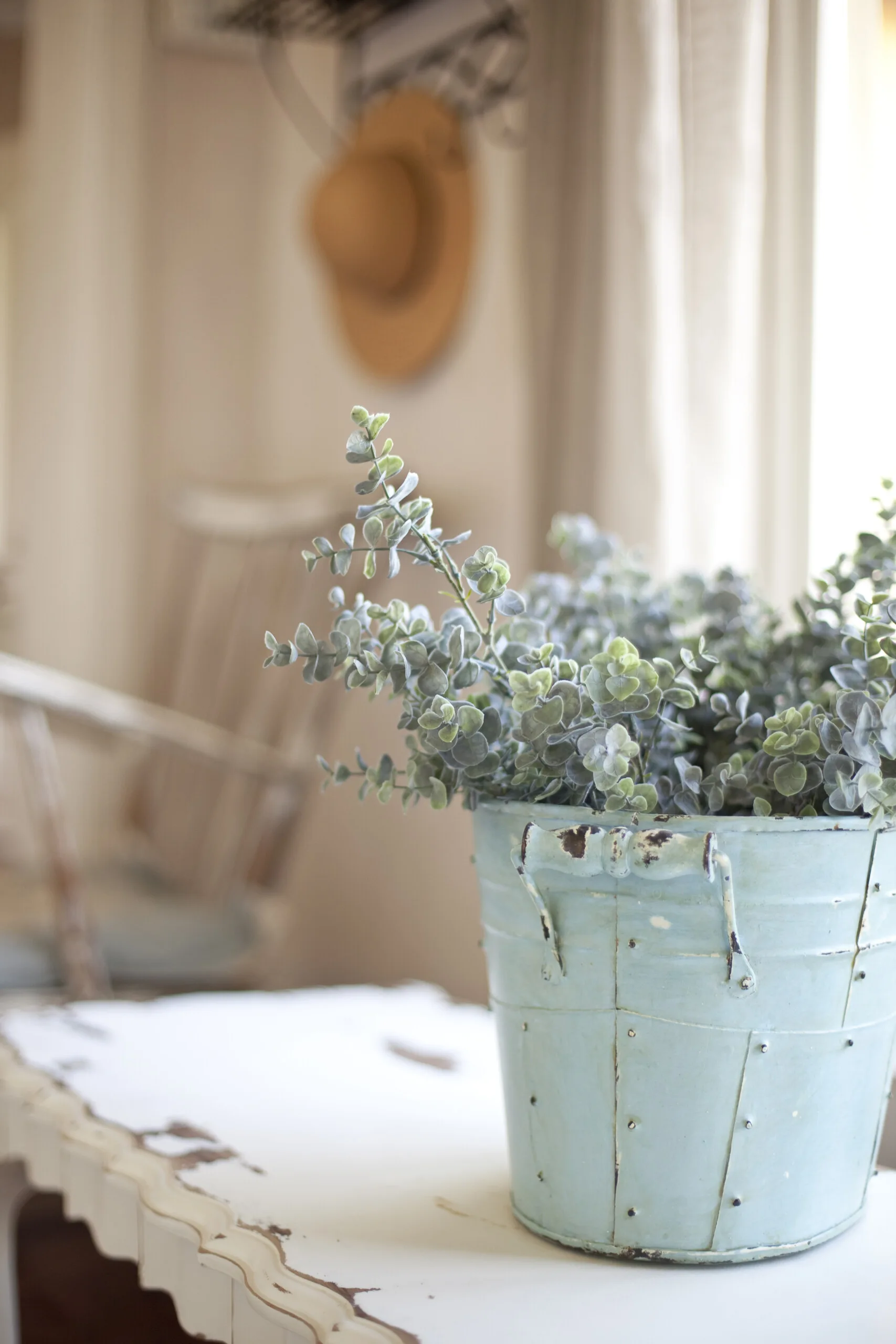 A distressed light blue metal bucket filled with green eucalyptus sits on a chipped white wooden table in front of a blurred background.