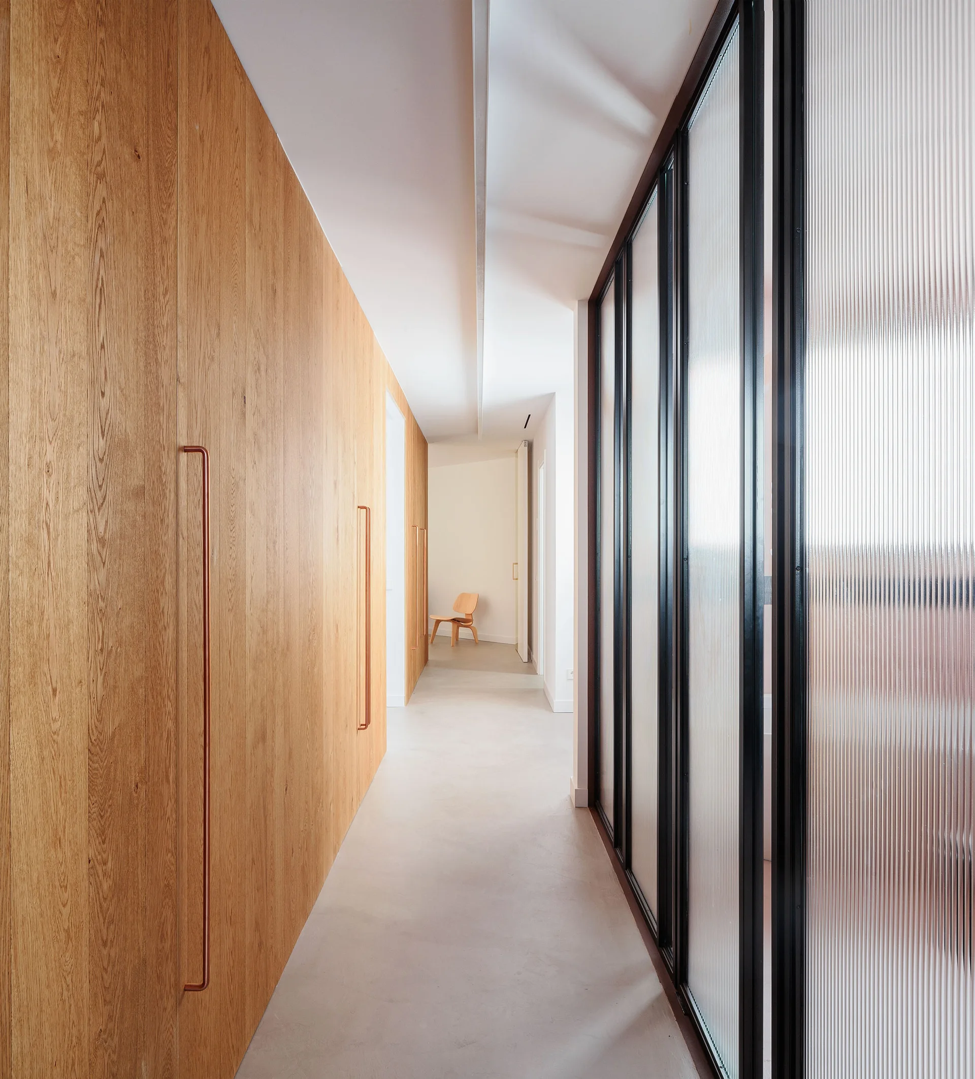 A minimalist hallway features seamless light wood wall paneling with copper handles opposite a black-framed fluted glass partition.