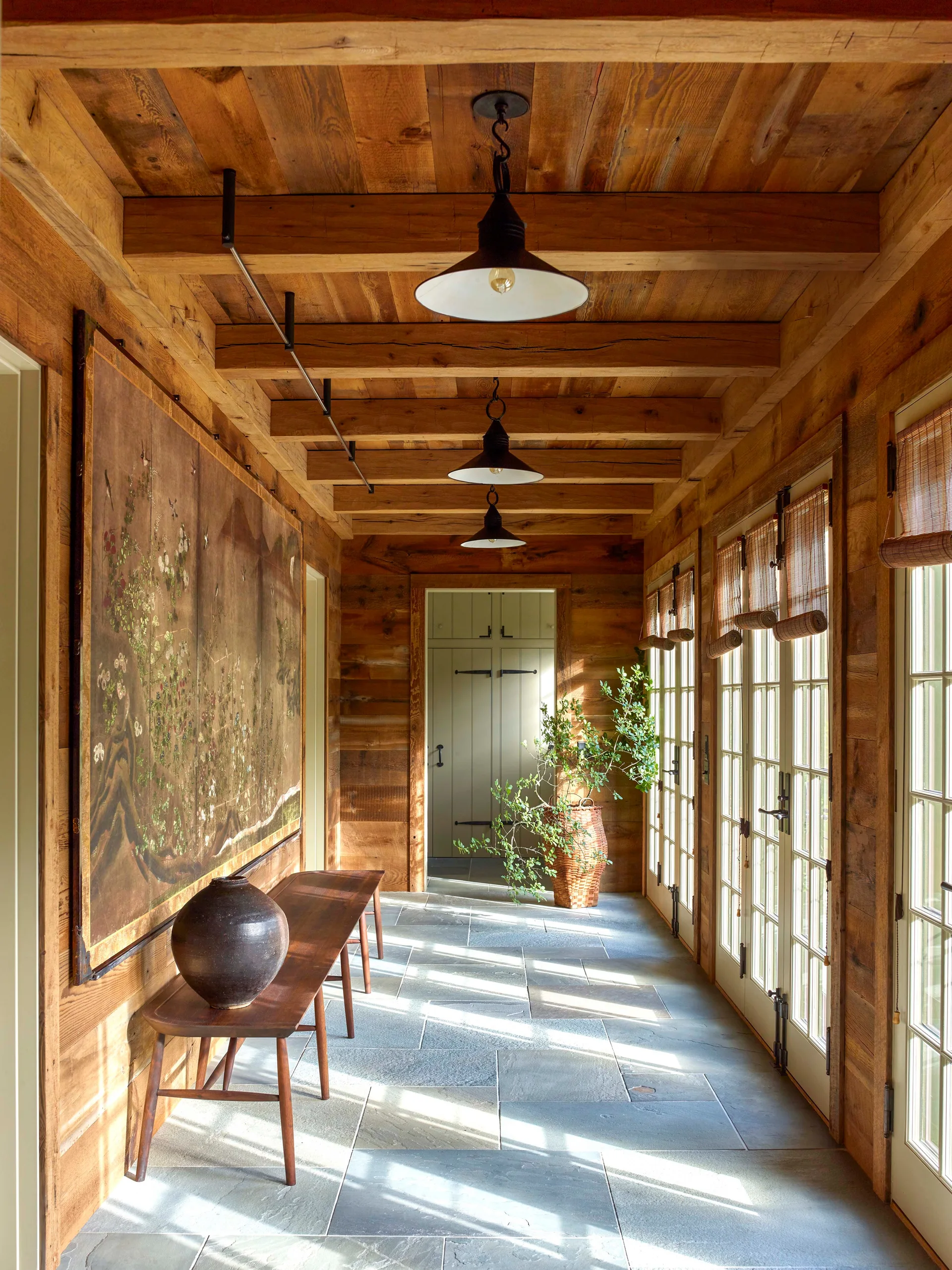 A rustic hallway features wood-paneled walls, exposed ceiling beams, flagstone tile flooring, a wooden bench, and glass french doors.