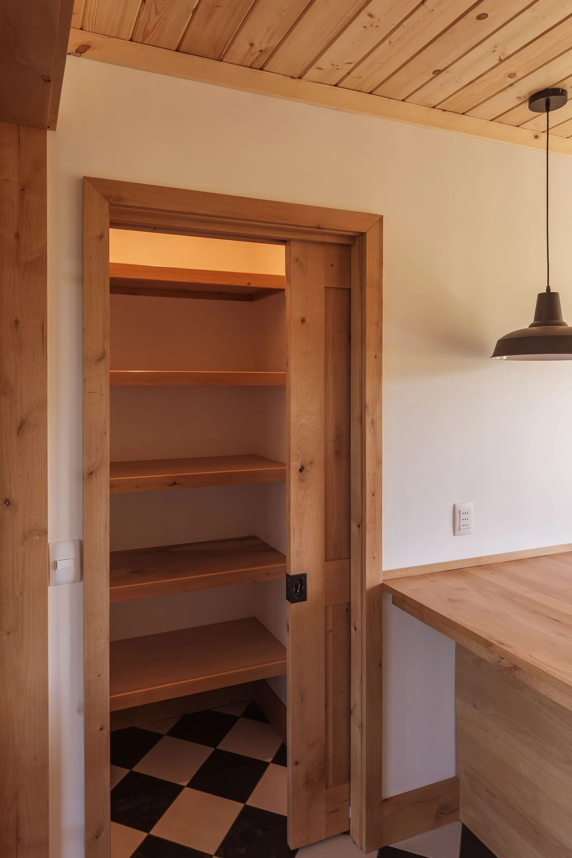 An open wooden pantry features a sliding pocket door, sturdy timber shelves, warm overhead lighting, and a black-and-white checkerboard tiled floor.
