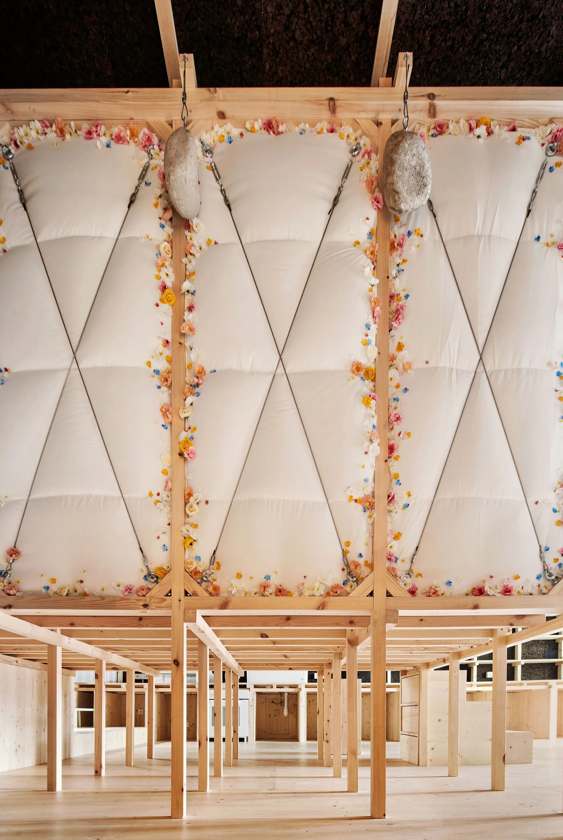 An overhead quilted white fabric canopy adorned with small colorful flowers is suspended above an unfinished light wood timber structure.