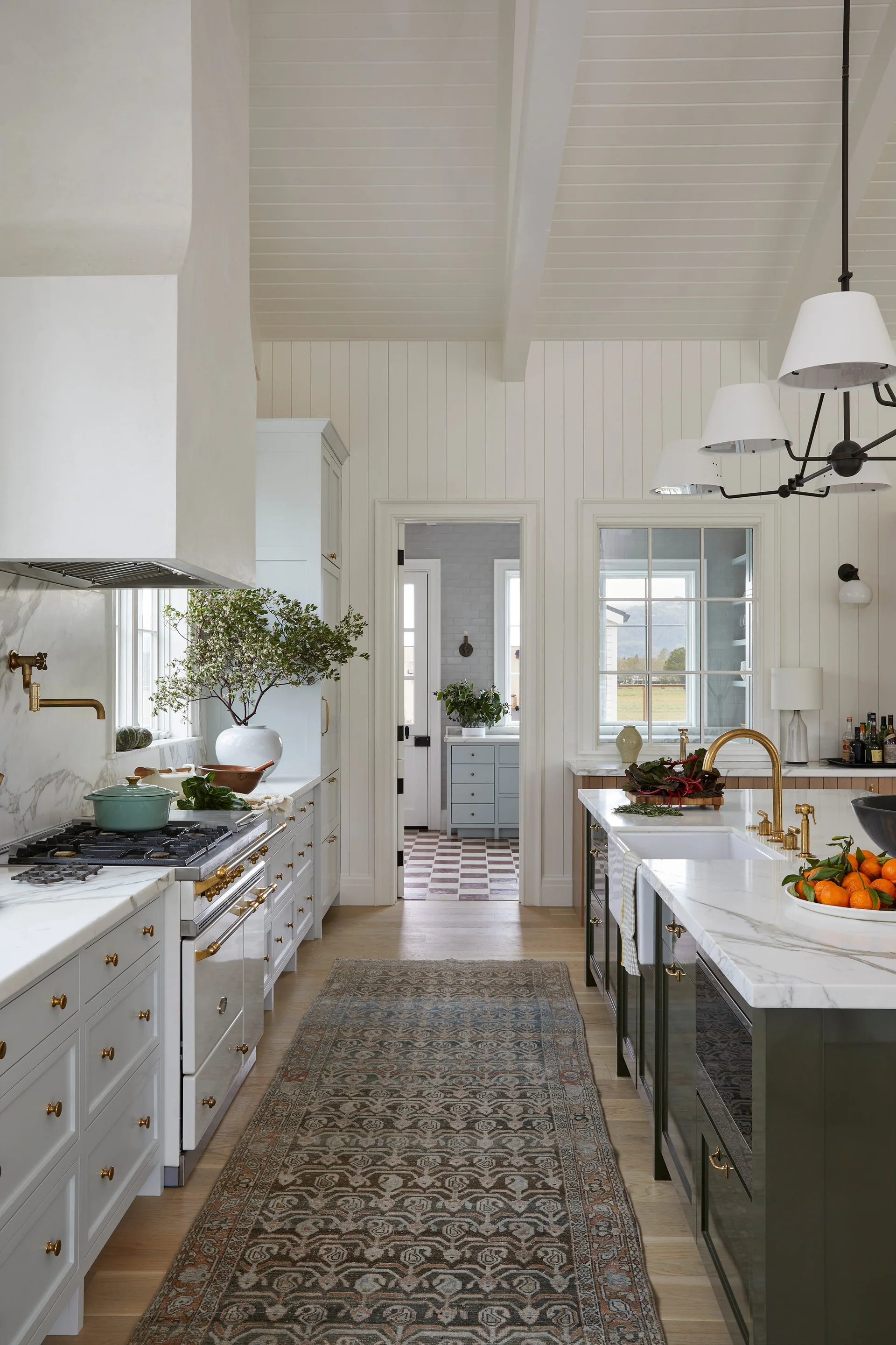 A bright farmhouse kitchen features white cabinetry and shiplap walls, a marble-topped island with a gold faucet, and a patterned runner rug.
