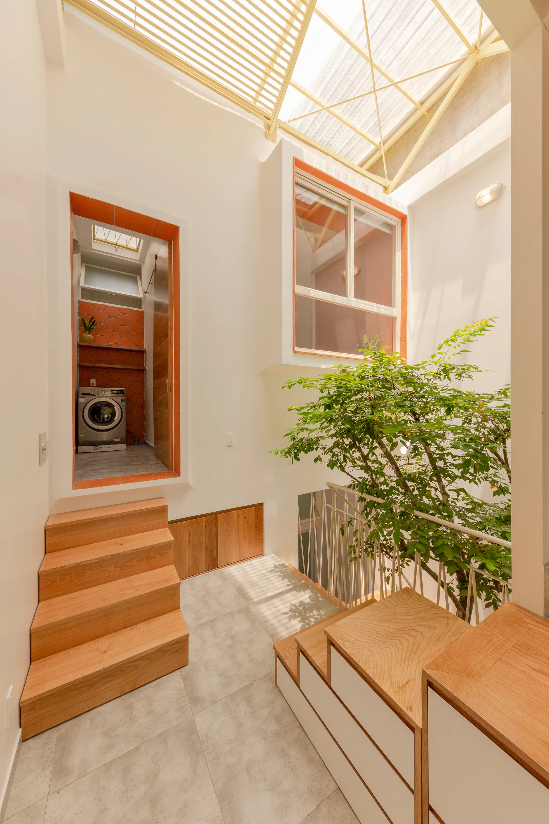 A light-filled interior atrium features a wooden staircase, a leafy indoor tree, and a doorway framed in terracotta tile leading to a laundry room.