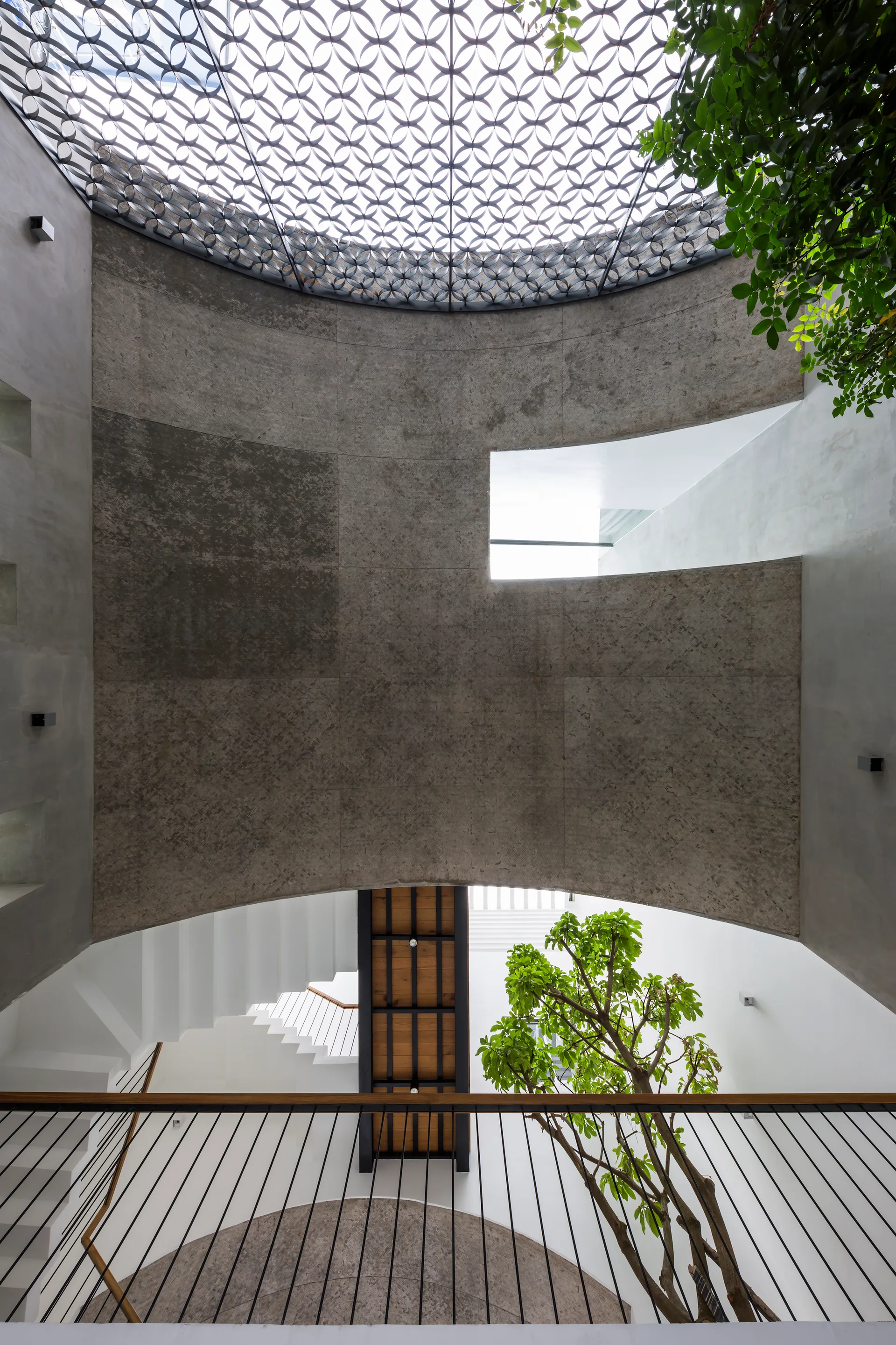 An upward view of a concrete atrium featuring a circular geometric lattice skylight, a white staircase, and an indoor tree.