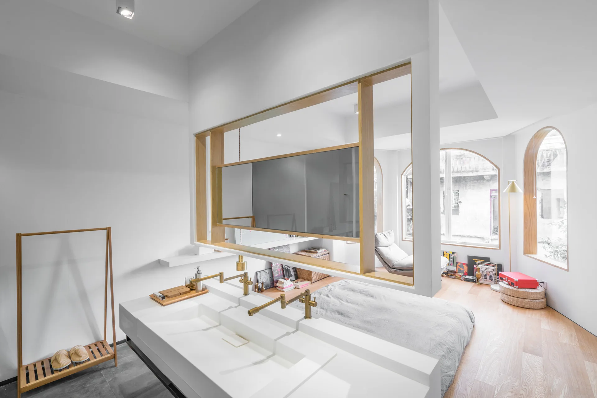 A white double vanity with brass faucets stands in front of a natural wood partition frame with a horizontal mirror, looking into an adjacent bedroom.