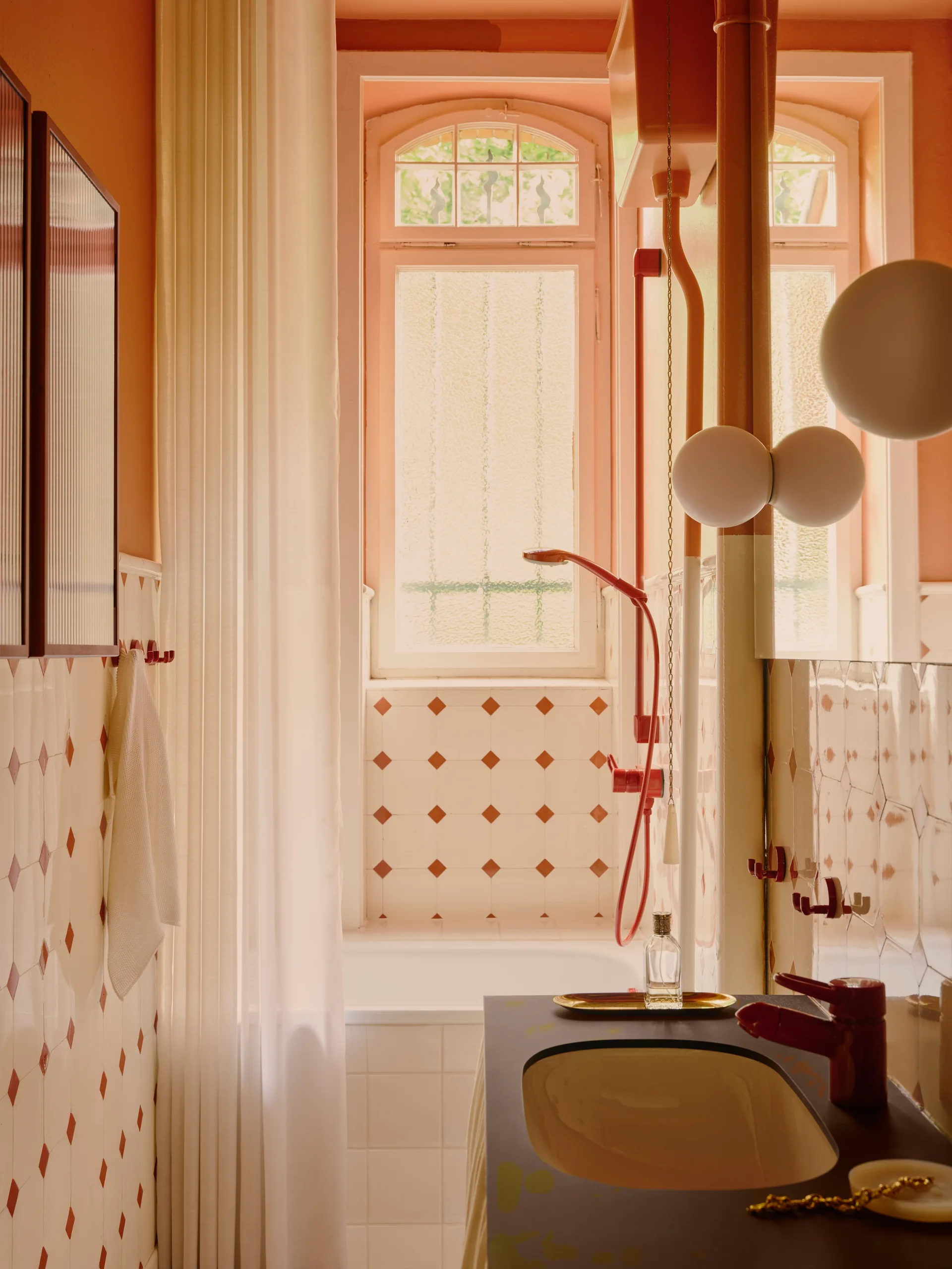A peach bathroom features a dark vanity, white tiles with red diamond accents, a white bathtub, and red fixtures beneath an arched window.