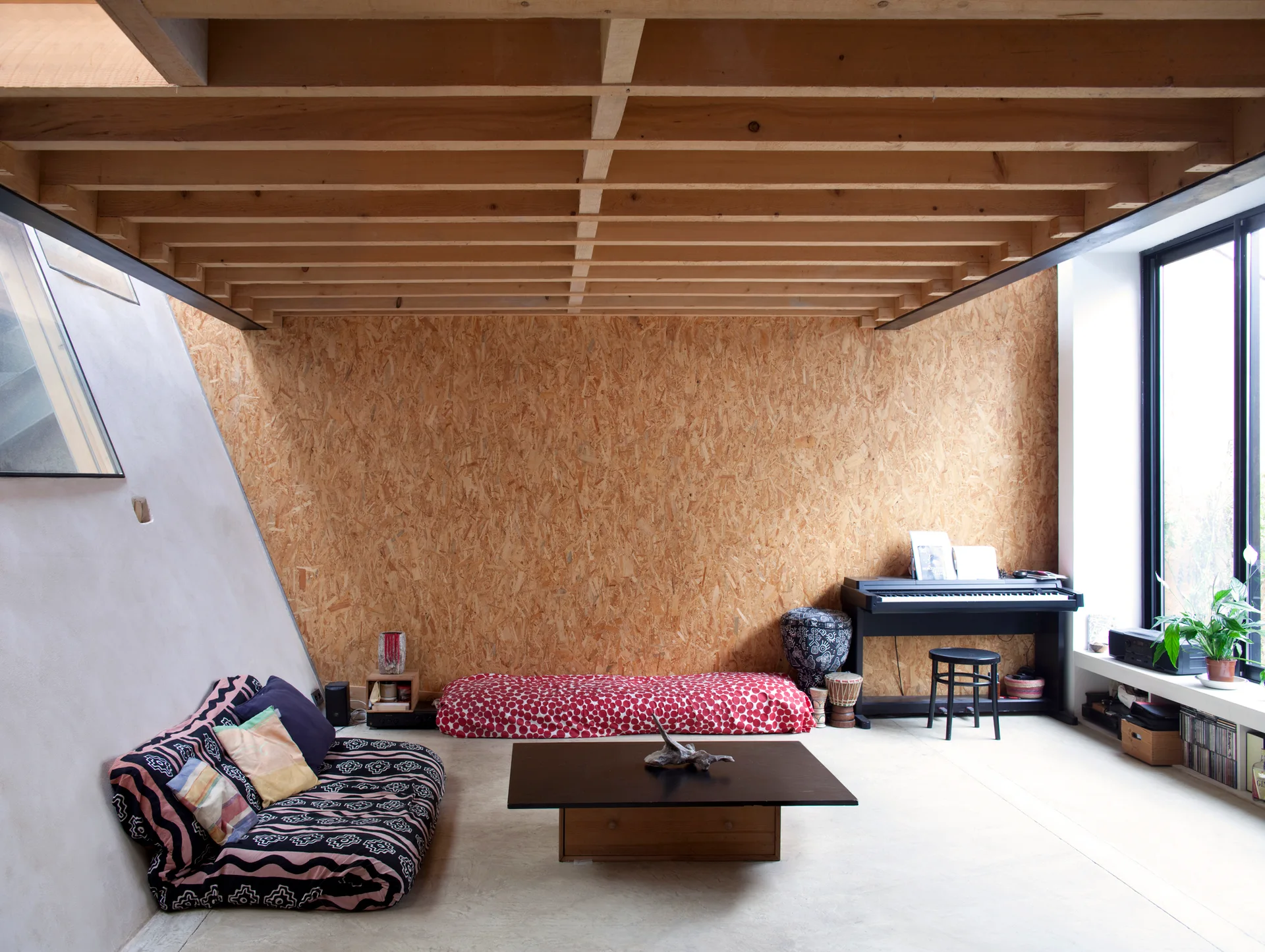 A casual living room features an oriented strand board wall, exposed timber ceiling joists, and low patterned floor cushions around a coffee table.
