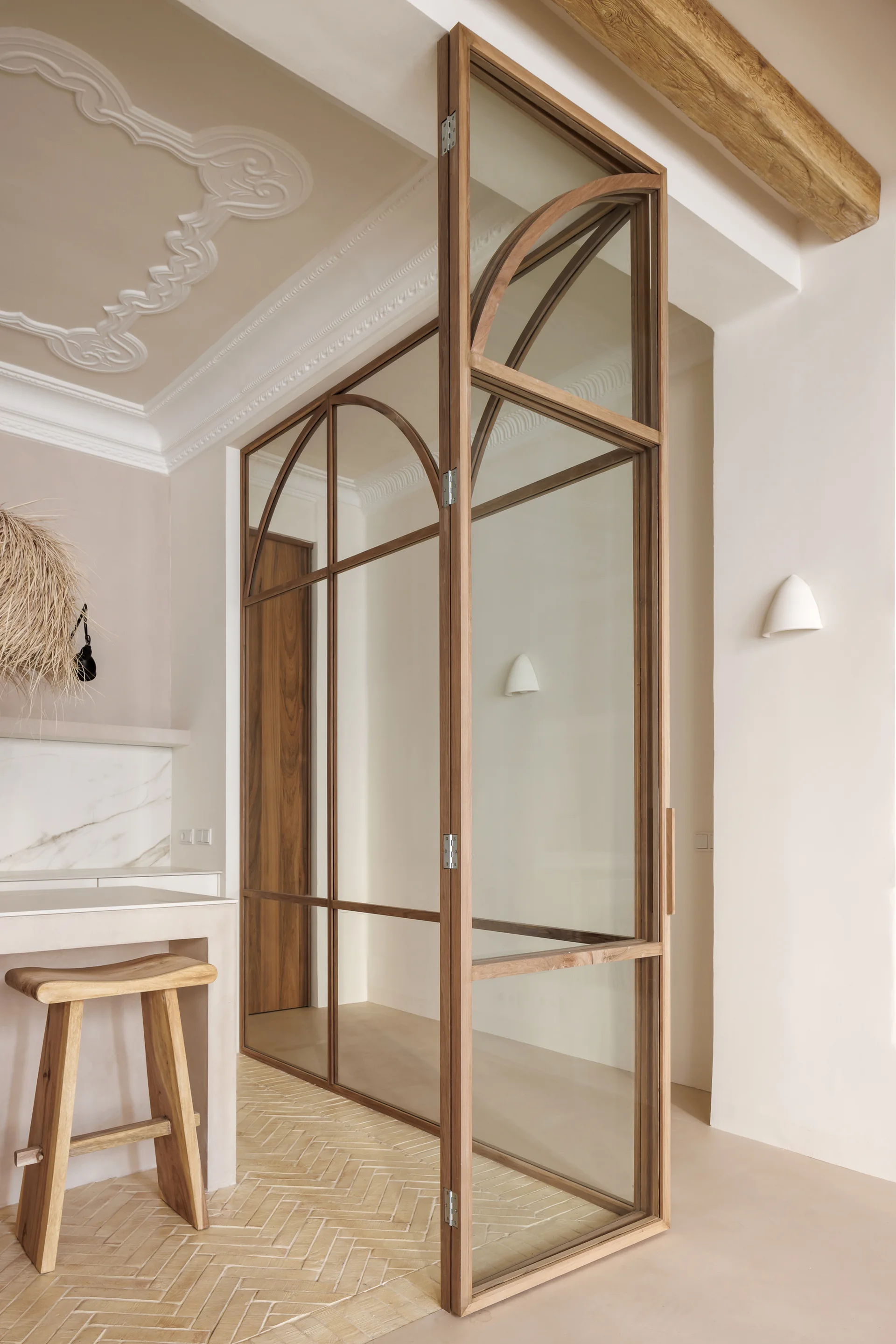 A tall wooden-framed glass partition with arched details stands next to a kitchen counter with a wooden stool and herringbone-patterned tile floor.