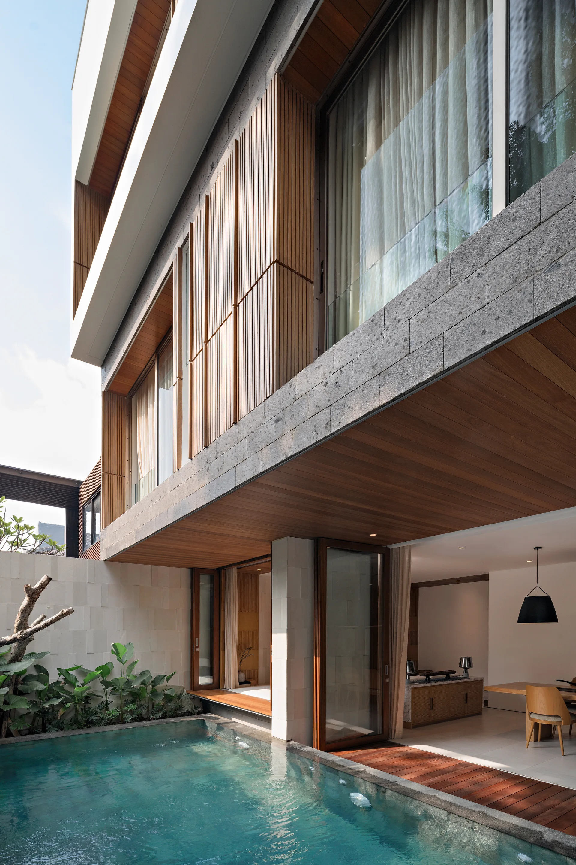 A modern multi-story home with a blue swimming pool next to an open-plan interior dining room, featuring wood ceilings and a stone facade.