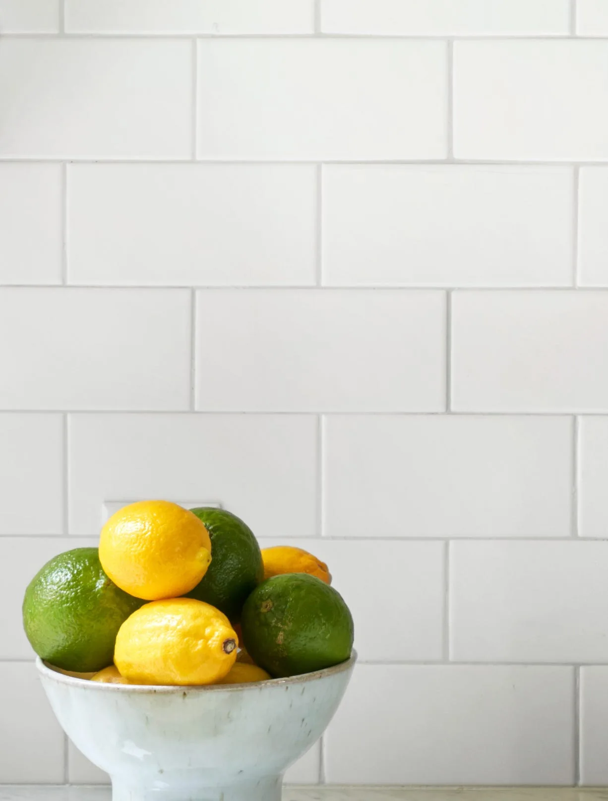 A pedestal ceramic bowl filled with green limes and yellow lemons stands against a white subway tile backsplash.