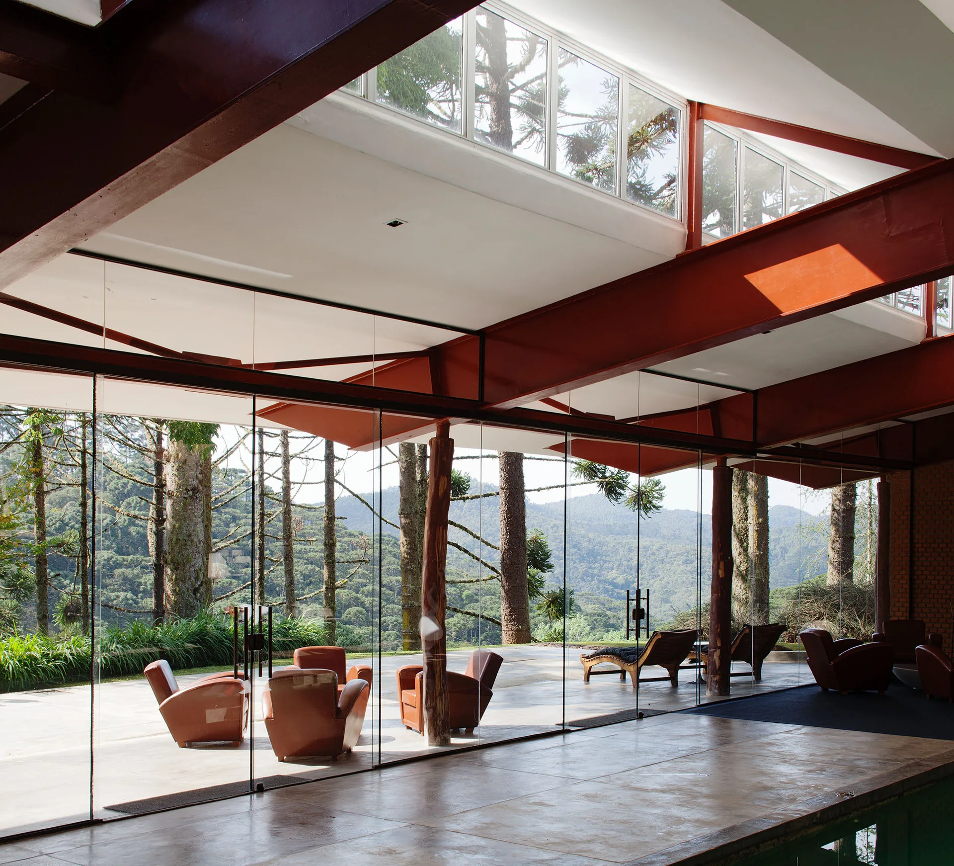 A modern living area with floor-to-ceiling glass walls, prominent red steel beams, natural log columns, and brown leather armchairs.
