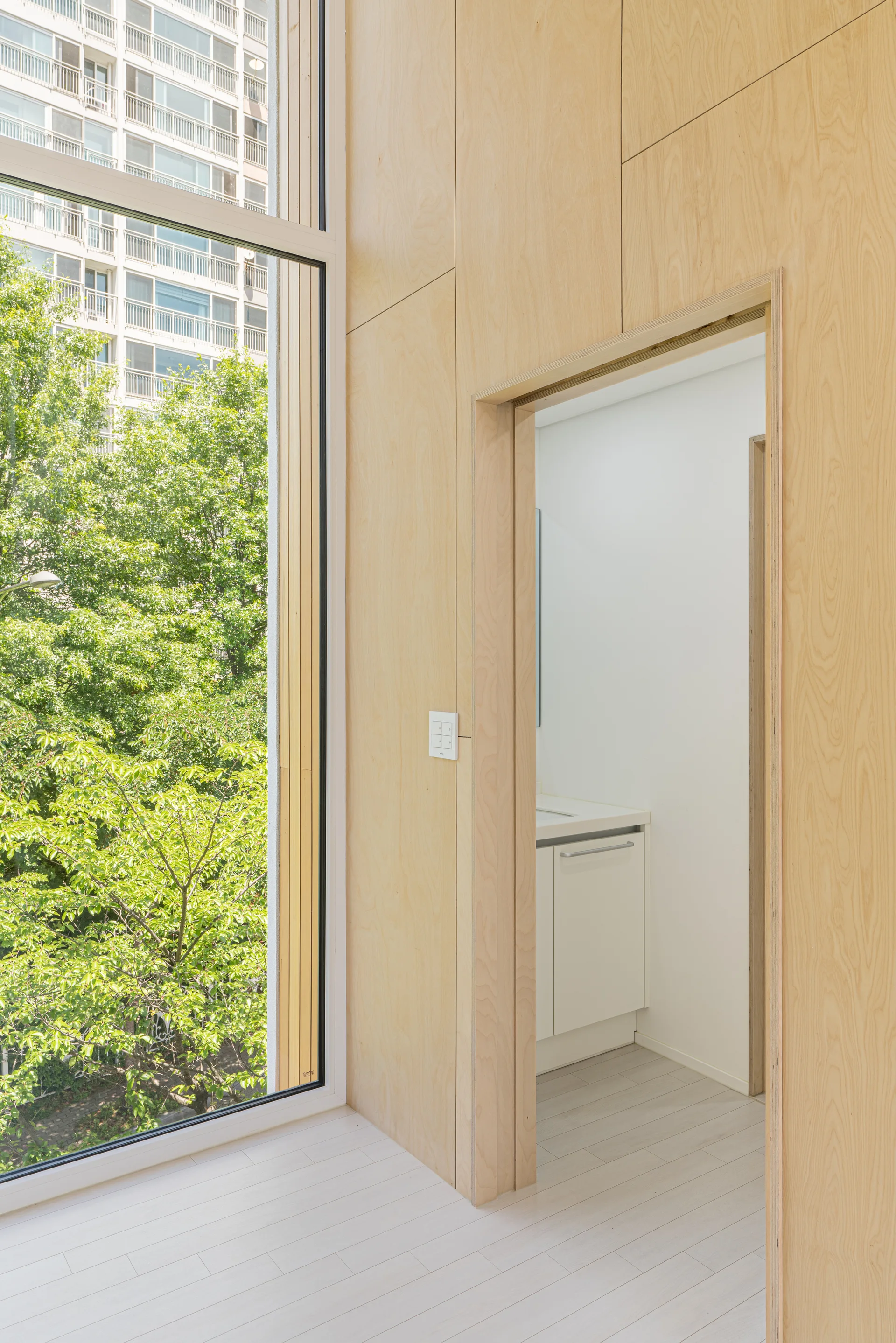 A light wood paneled hallway features a tall window looking out to green trees and a doorway leading into a white bathroom with a vanity.