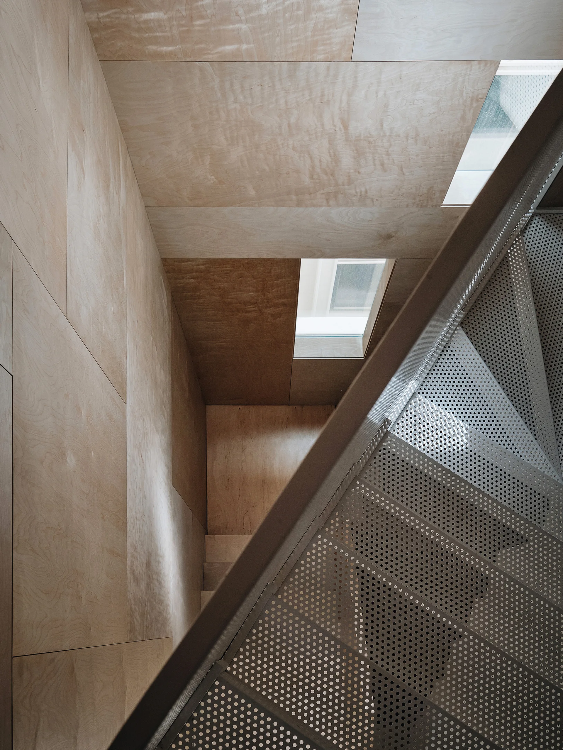 A high-angle view looking down a stairwell with perforated metal steps, light plywood-paneled walls, and a small recessed window.
