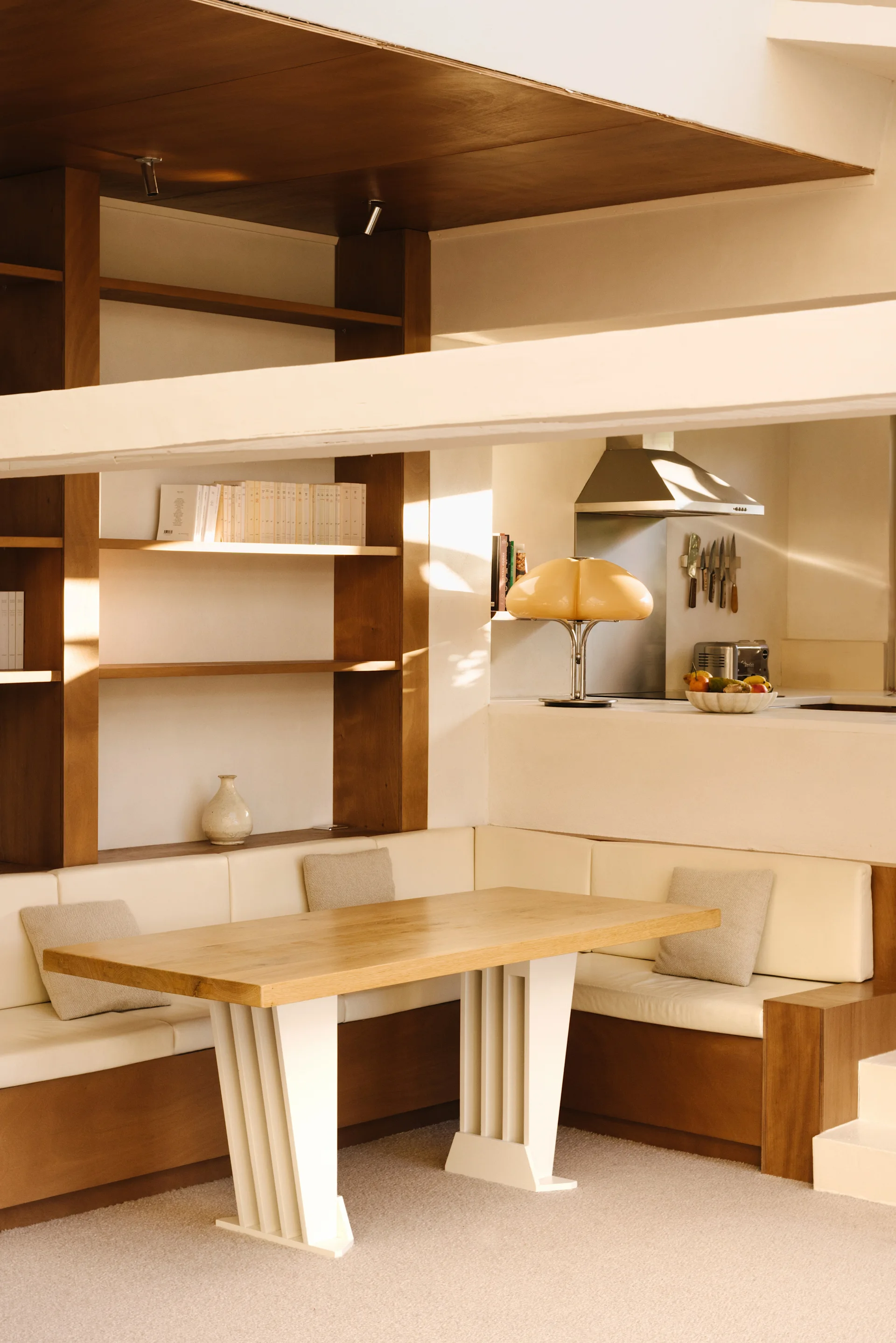 A built-in dining nook features a warm wood table, cream banquette seating, and integrated shelves next to a kitchen counter with an amber lamp.
