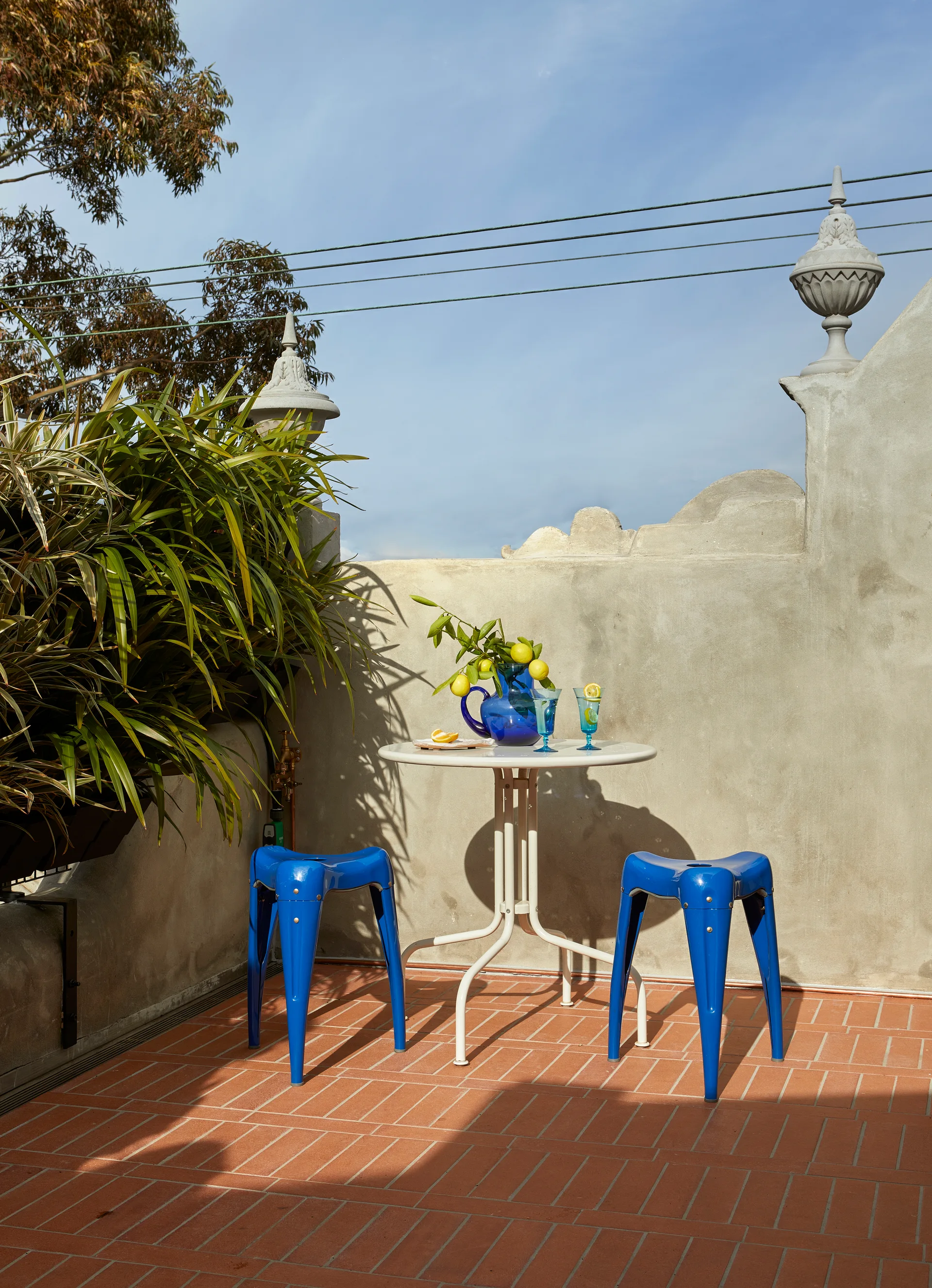 A sunny outdoor patio features a white metal bistro table and two glossy blue stools on a terracotta tile floor against a grey concrete wall.