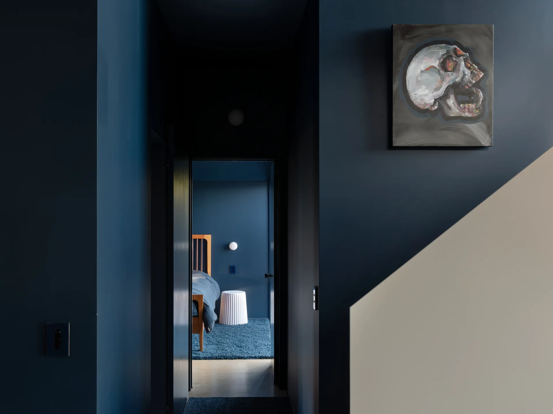 A dark navy blue hallway with a skull painting leads into a bedroom with a wooden bed frame, a white fluted side table, and a blue rug.