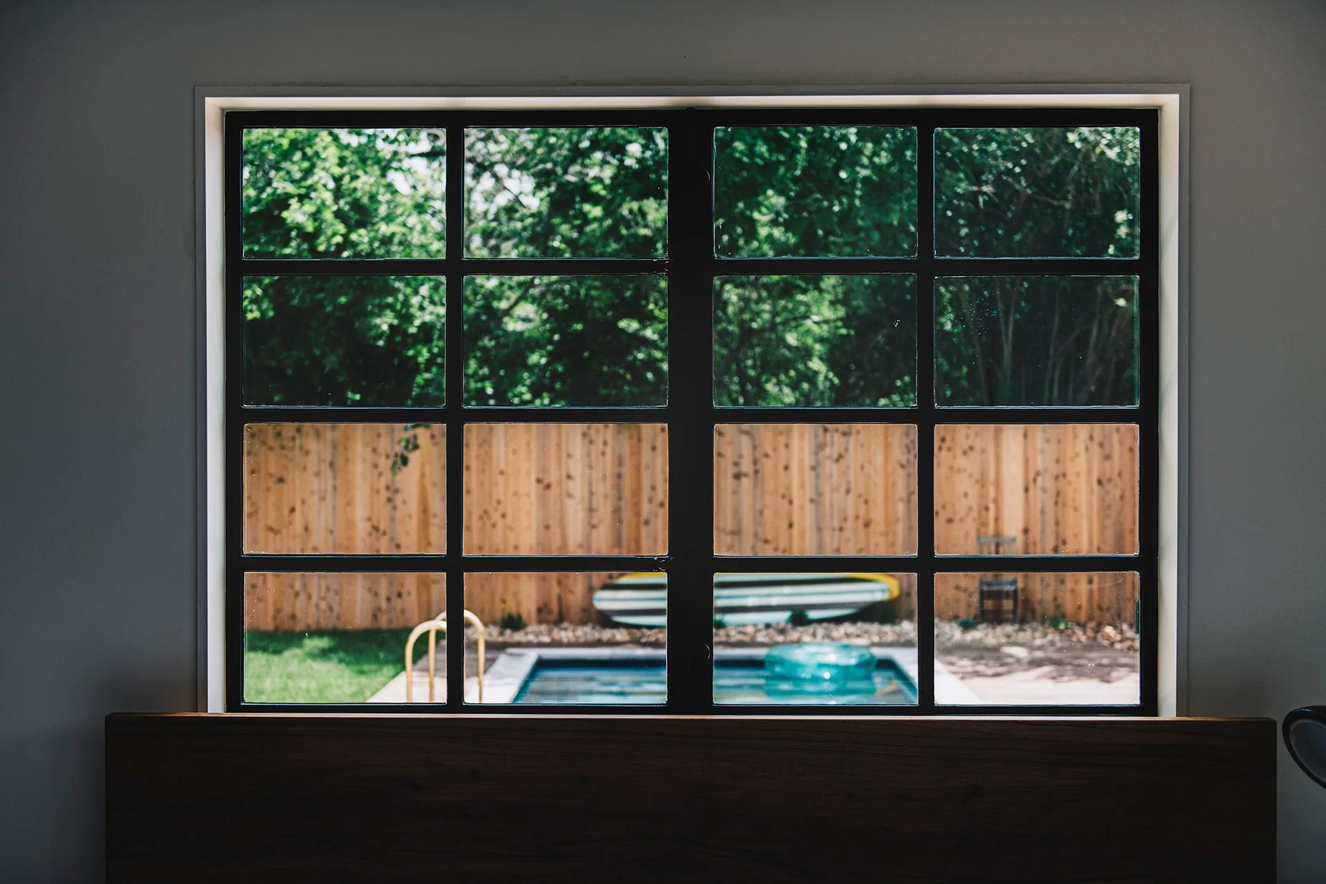 A black-framed multi-pane window overlooks a backyard swimming pool with a wooden fence and green trees.
