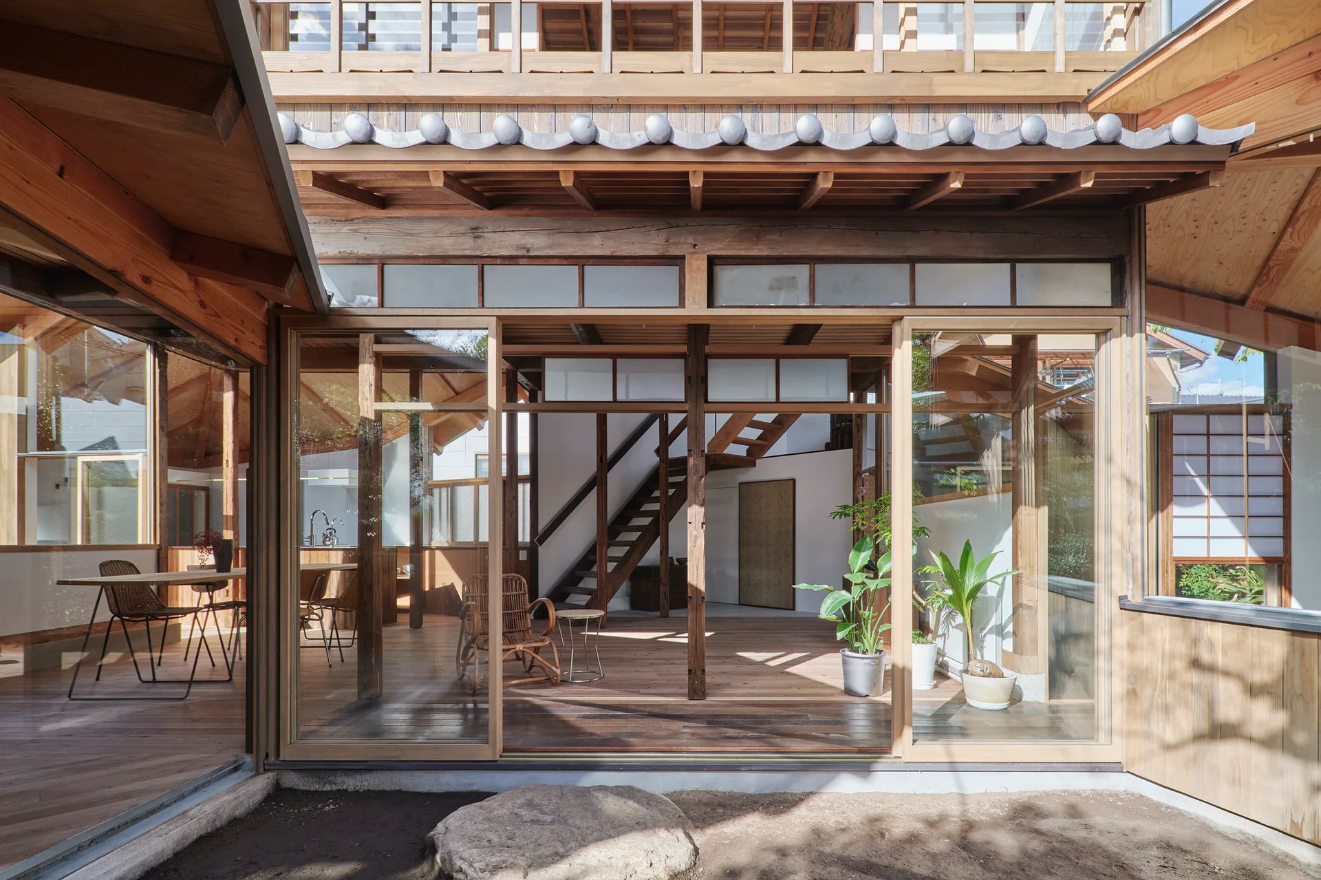 A modern Japanese-inspired home features large sliding glass doors, exposed timber framing, a central wooden staircase, and a minimal living area.