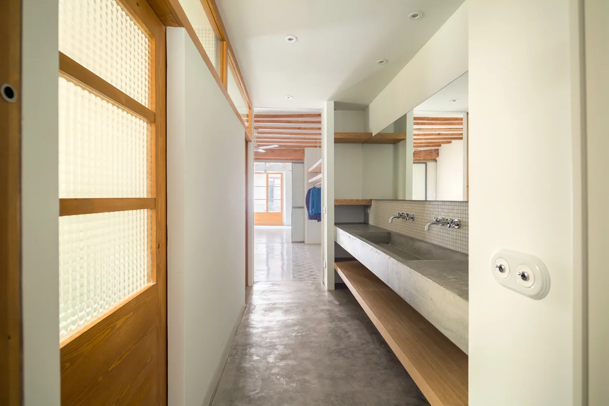 A modern bathroom hallway featuring a long concrete double vanity sink, wall-mounted fixtures, and a wooden shelf.