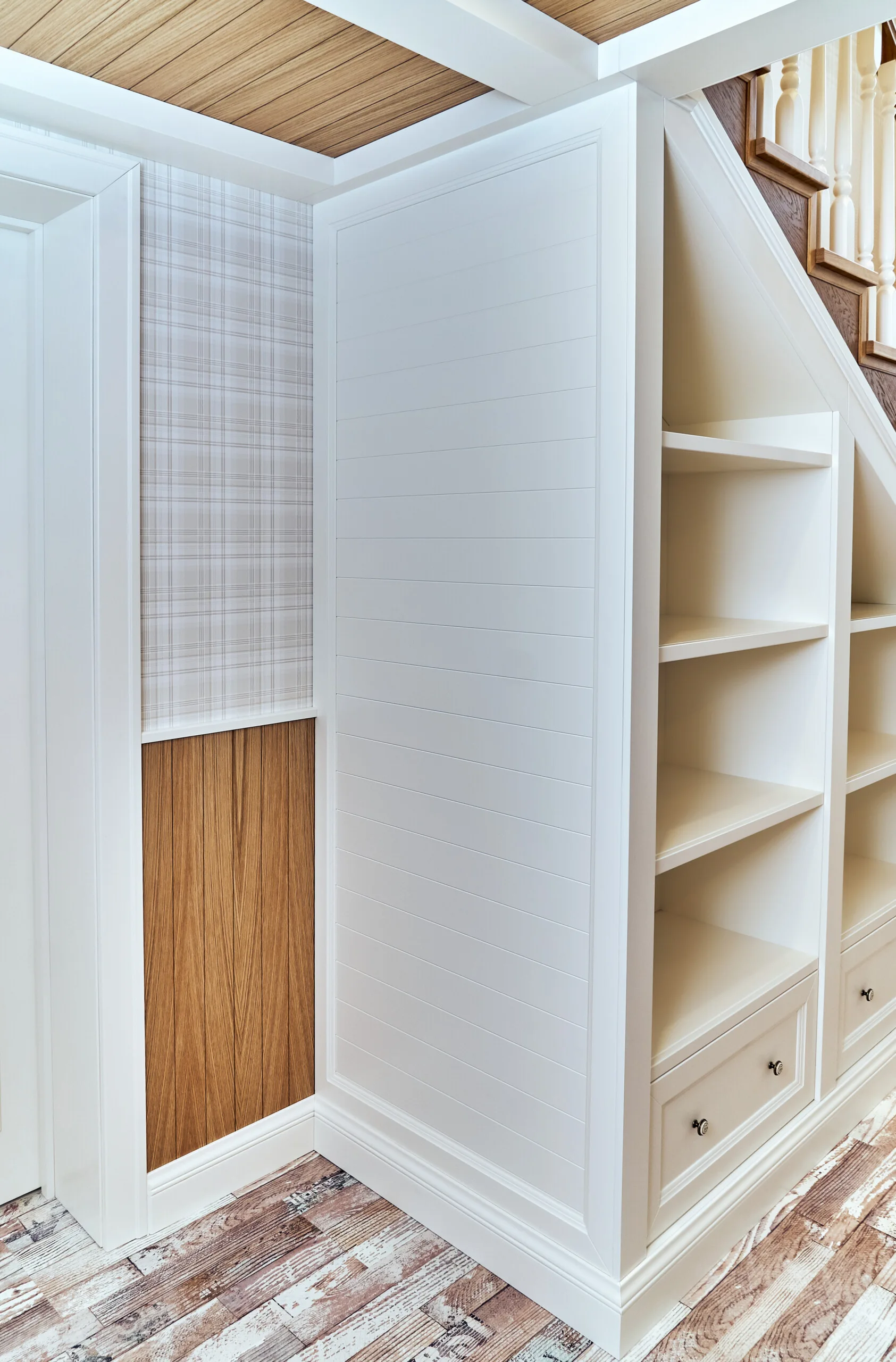 A white built-in storage unit with open shelving and drawers is installed under a staircase next to plaid wallpaper.