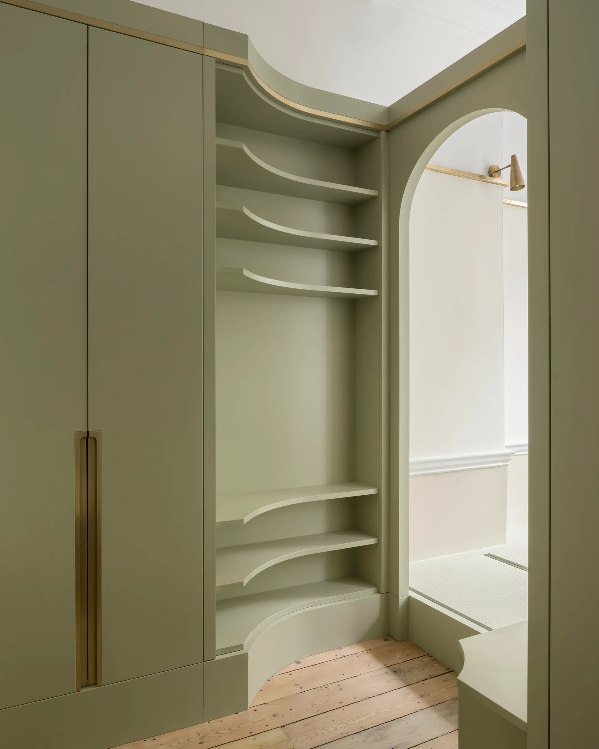 Custom sage green cabinetry features built-in curved shelves, a recessed brass handle, and an adjacent arched doorway over wood plank floors.