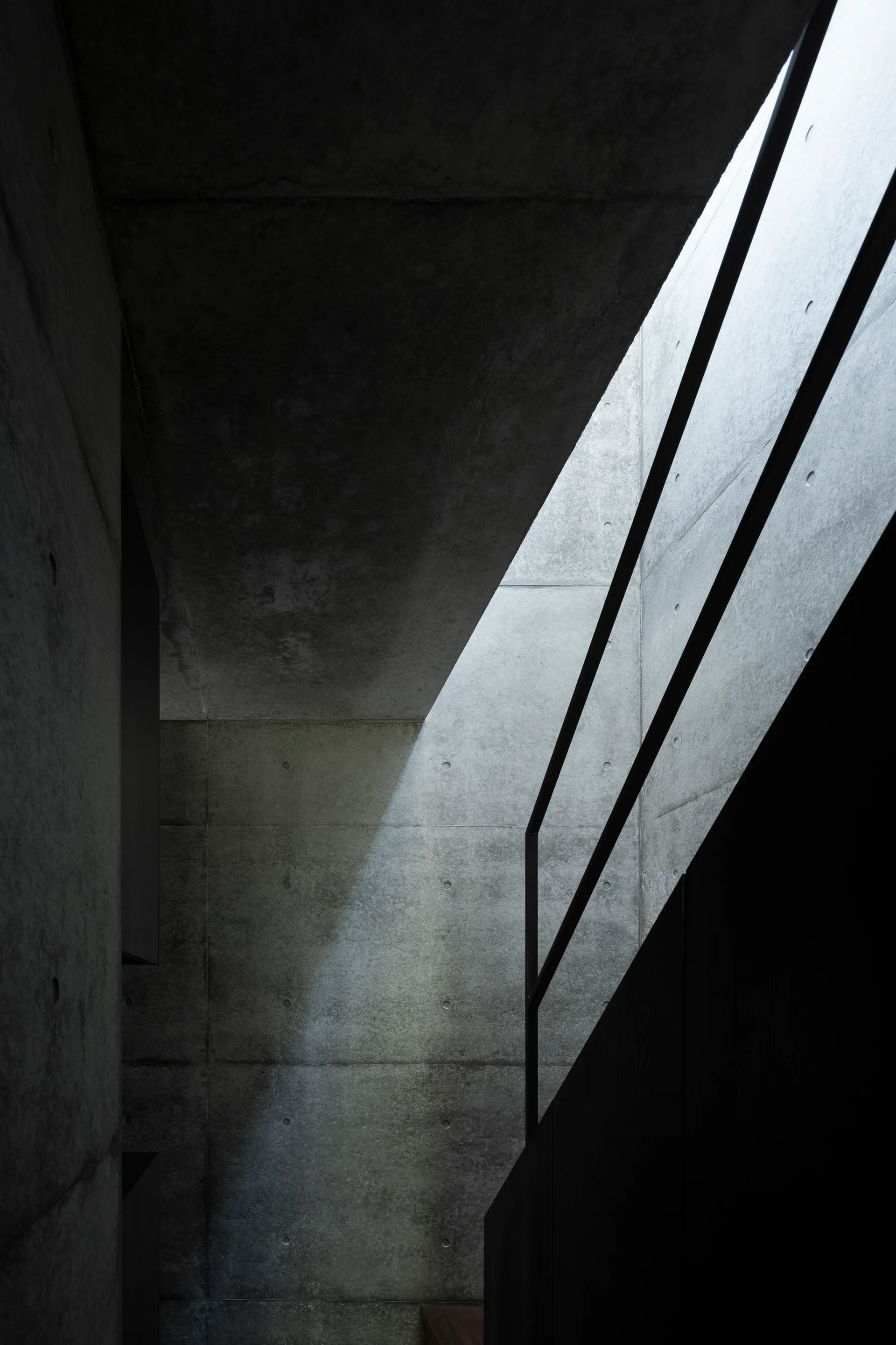 A minimalist concrete stairwell features a slim black metal railing and natural light streaming diagonally from an overhead skylight.