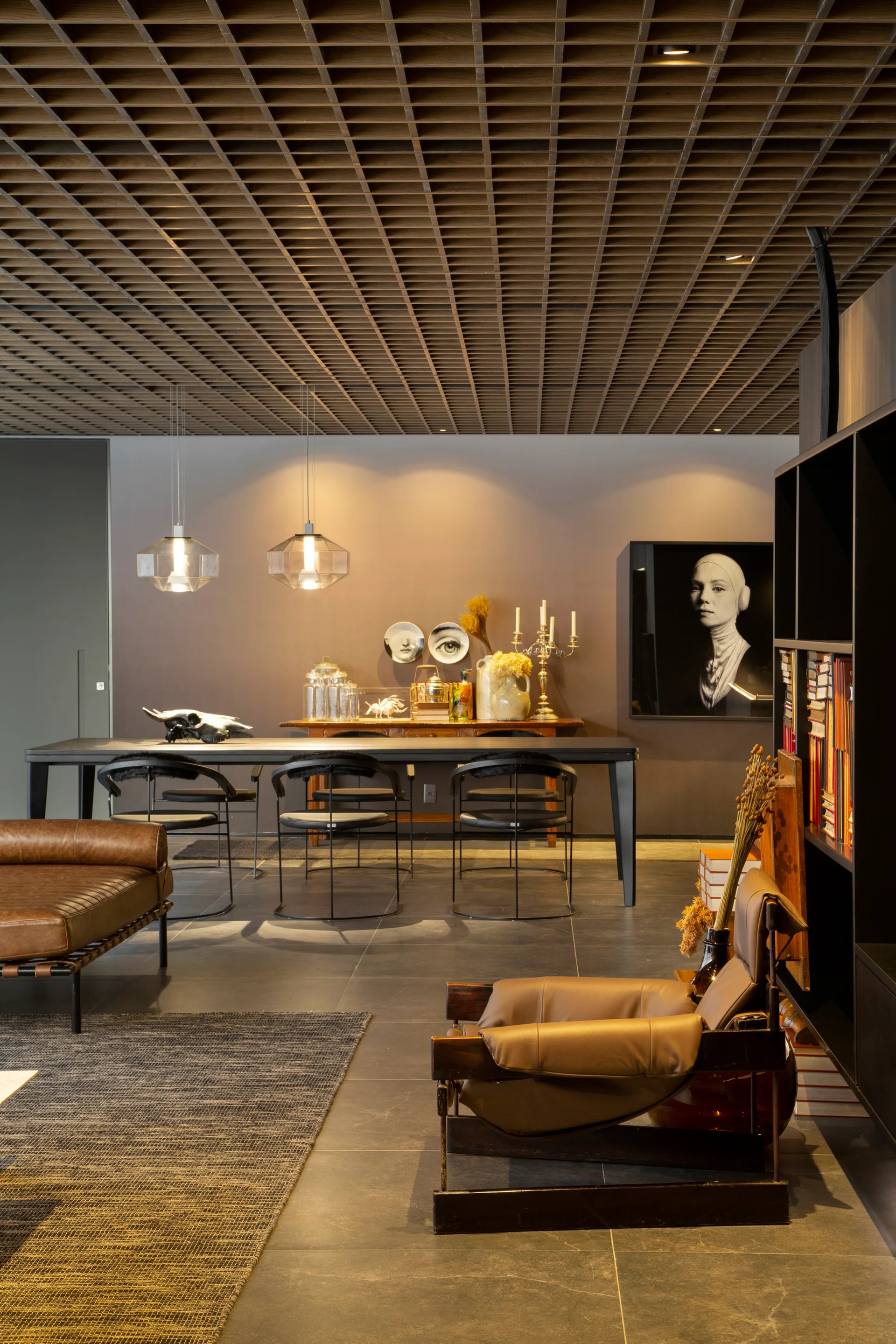 A contemporary living and dining area with a distinctive wooden grid ceiling, leather armchairs, a long black dining table, and dark tile flooring.