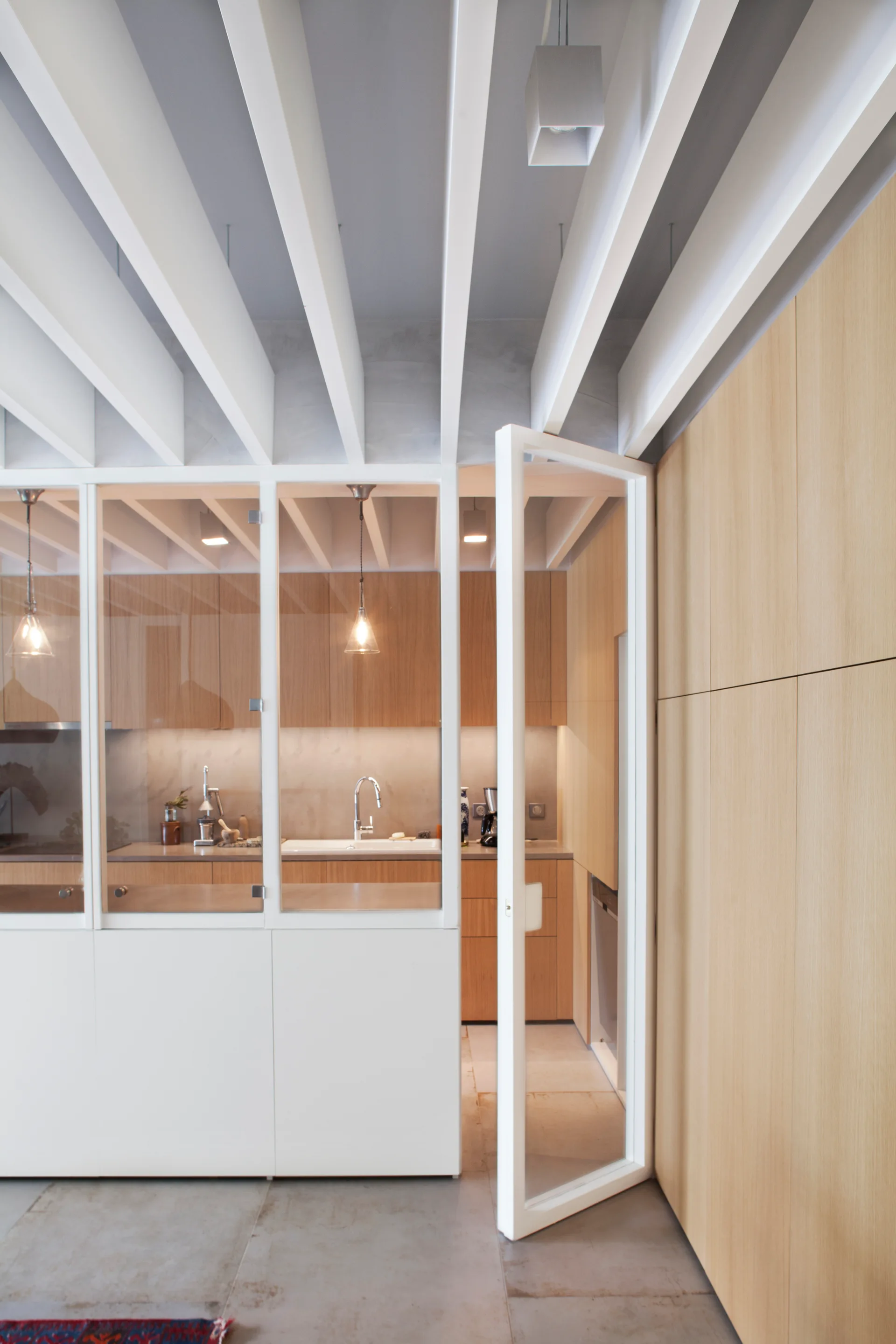 A modern kitchen with light wood cabinetry is visible through a white-framed glass partition wall beneath dramatic white exposed ceiling beams.