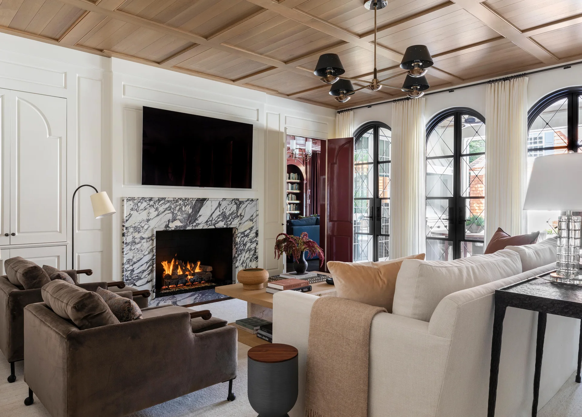 A living room featuring a wood coffered ceiling, a heavily veined marble fireplace, arched black glass doors, and a cream sofa.