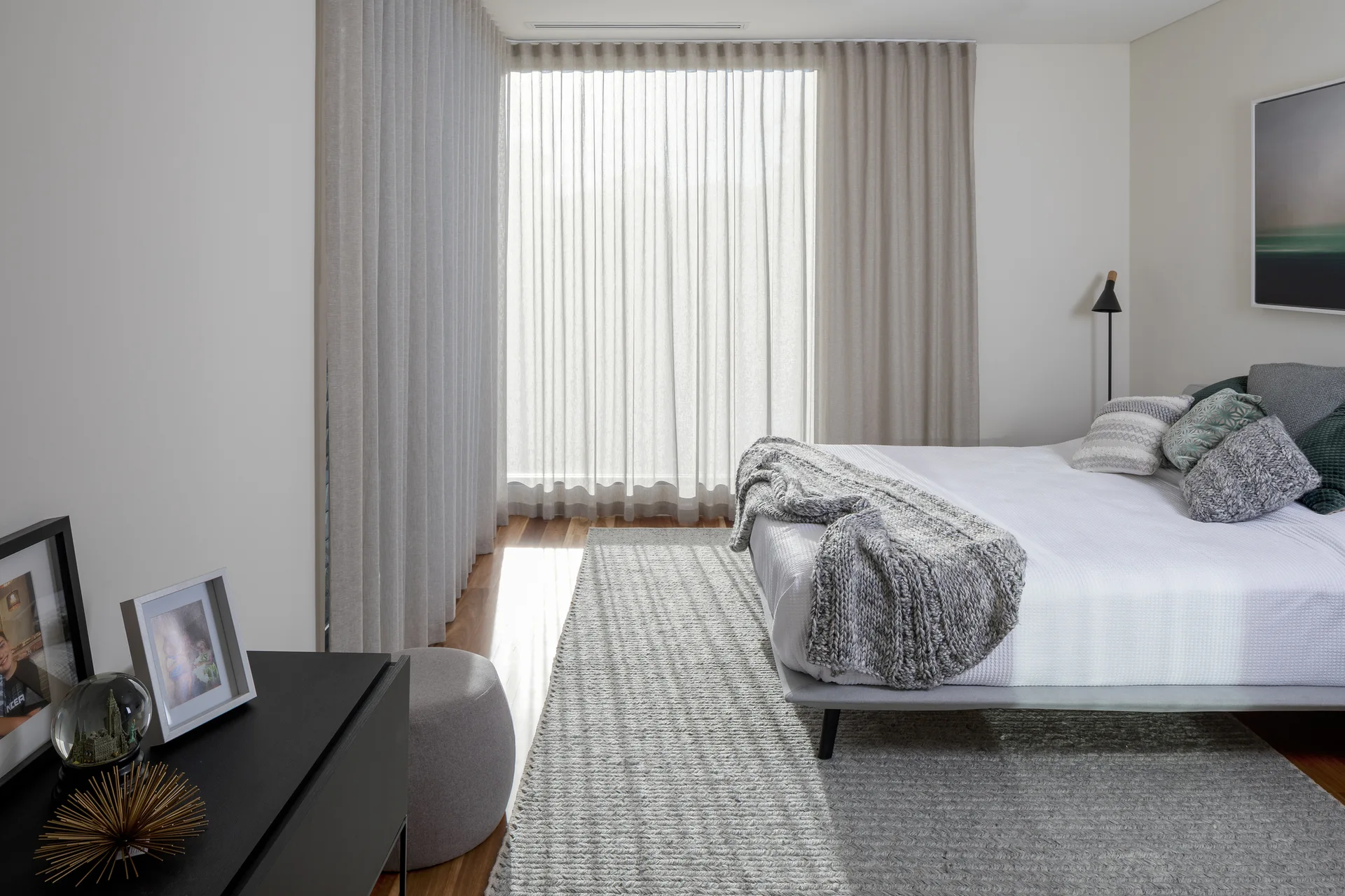 A minimalist bedroom features a gray textured area rug, a low bed with a chunky knit throw, and floor-to-ceiling sheer curtains.