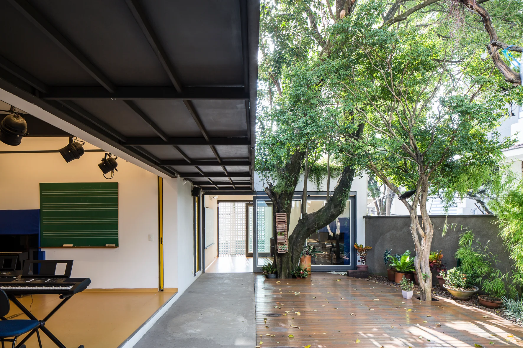 An indoor-outdoor music studio features a black metal ceiling structure, a green chalkboard, and a wooden deck with a large mature tree.