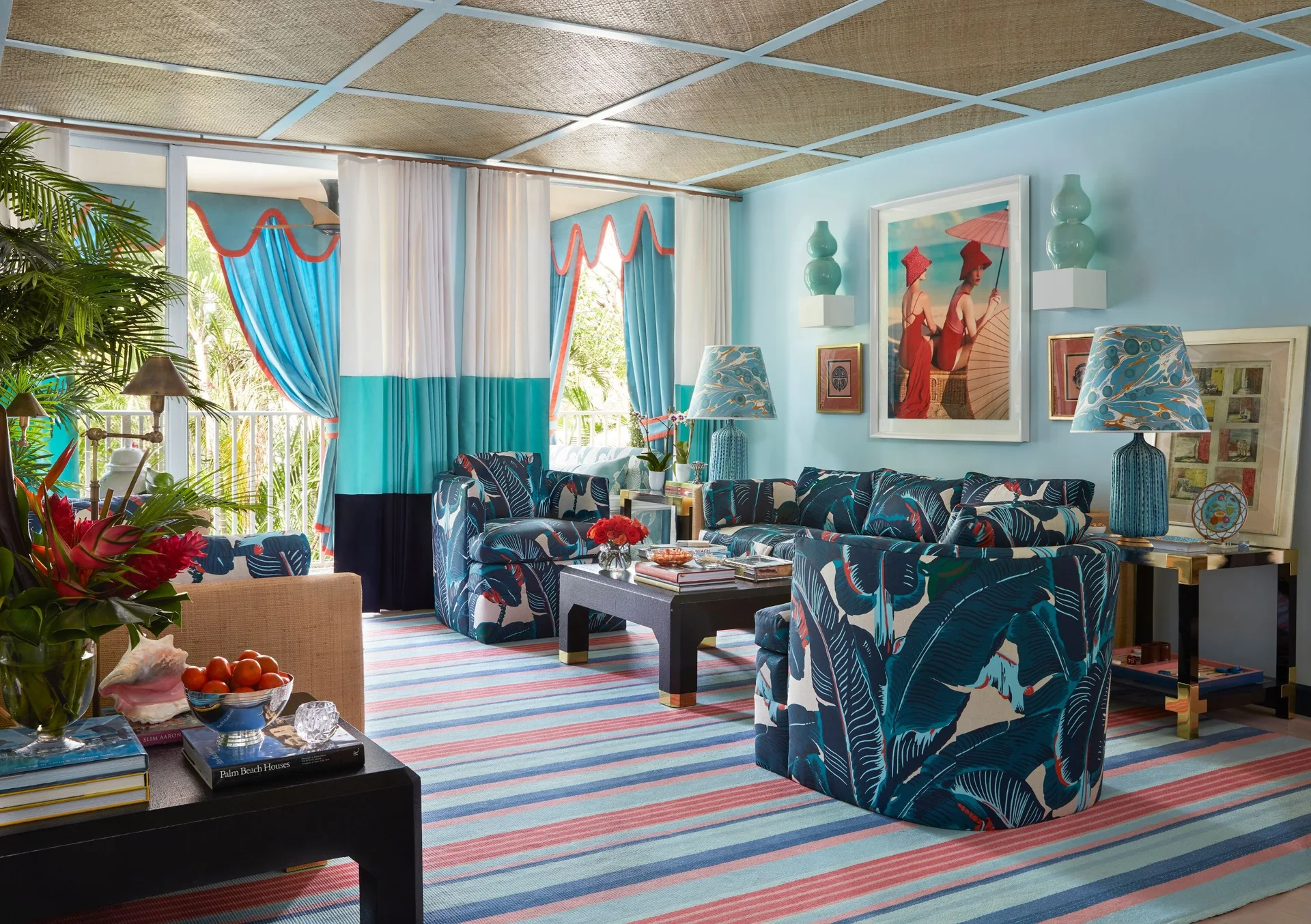 A light blue living room features tropical leaf patterned sofas, a striped area rug, and a textured woven grid ceiling with dark wood tables.
