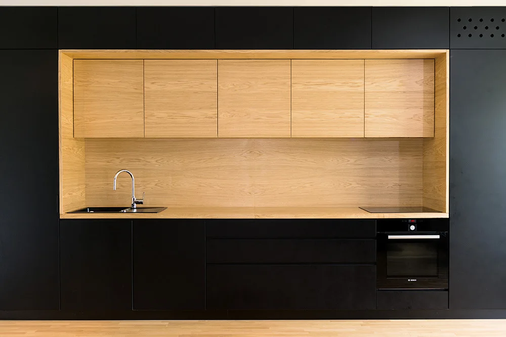 Minimalist matte black kitchen cabinetry surrounding a central recessed prep area lined with light oak panels and an integrated sink.