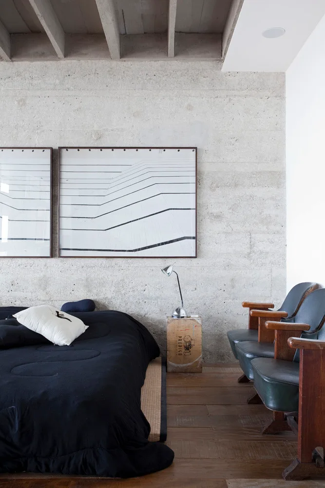 An industrial bedroom features a black low-profile bed, an exposed concrete block wall, a wooden crate nightstand, and two vintage theater chairs.