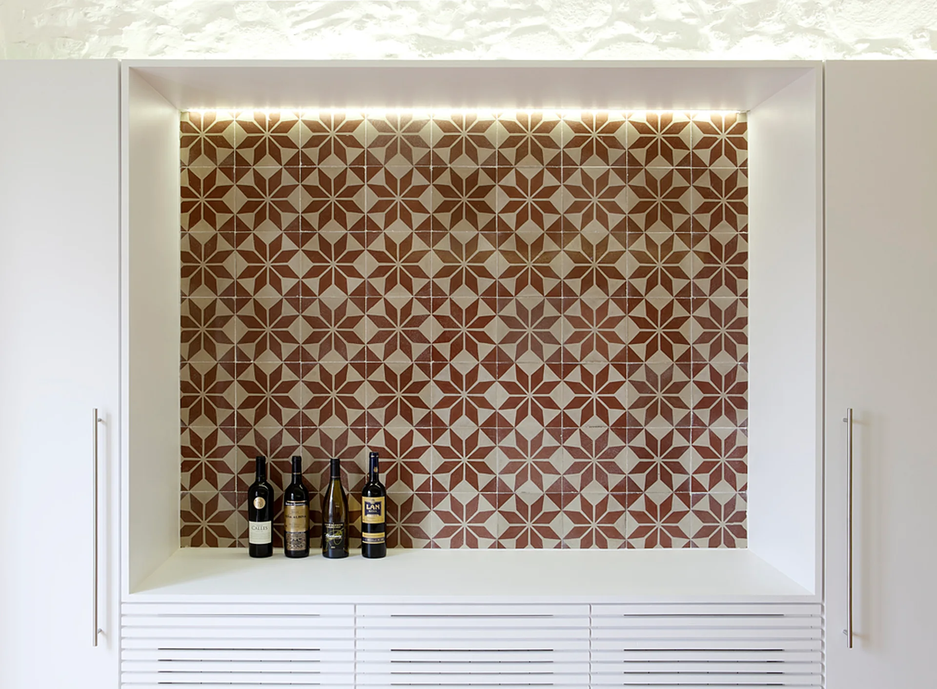 A built-in white bar cabinet features a star-patterned terracotta tile backsplash illuminated by overhead lighting with wine bottles on the counter.