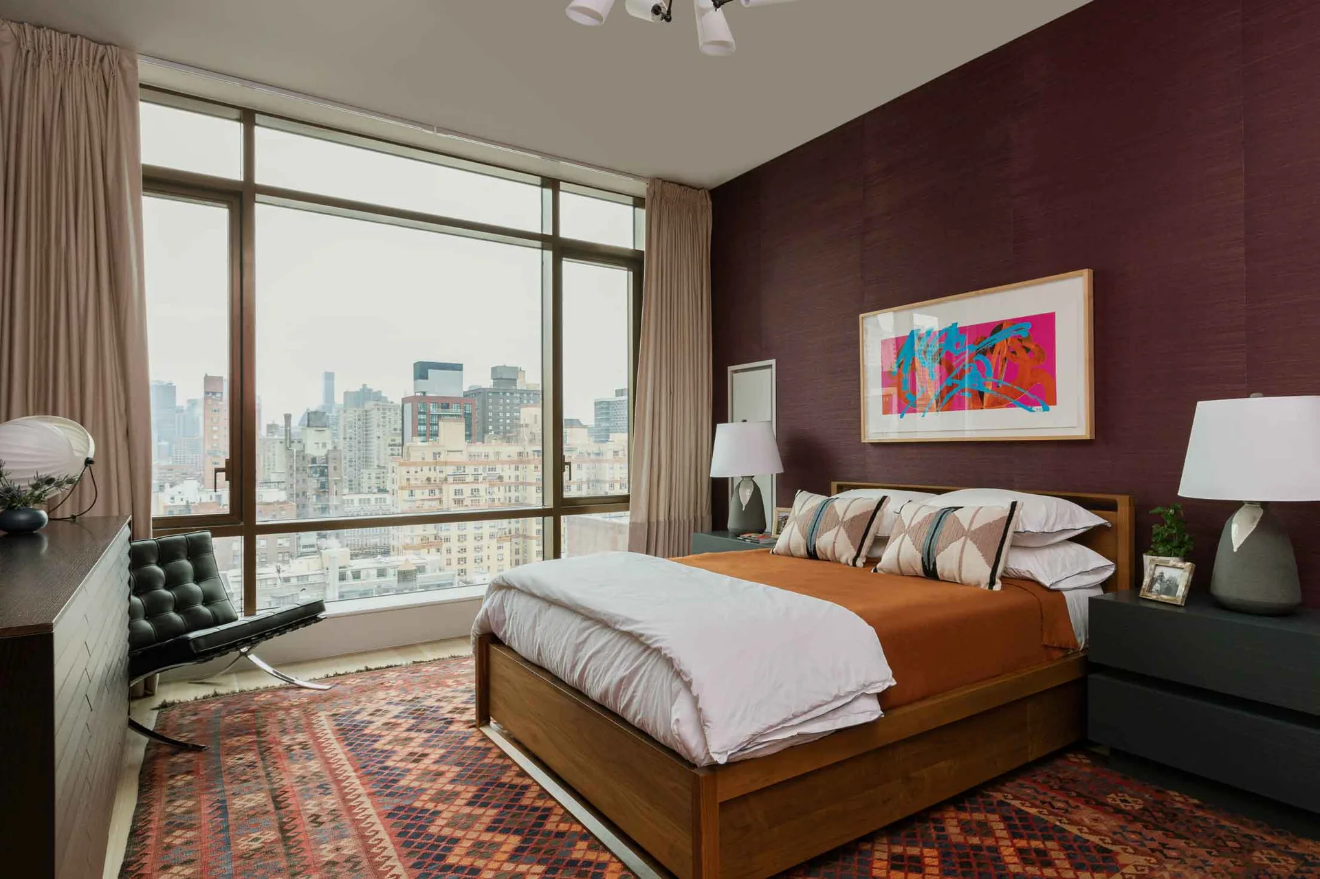 A modern bedroom features a wooden bed frame against a textured maroon wall, a colorful art piece, and large windows overlooking a city skyline.